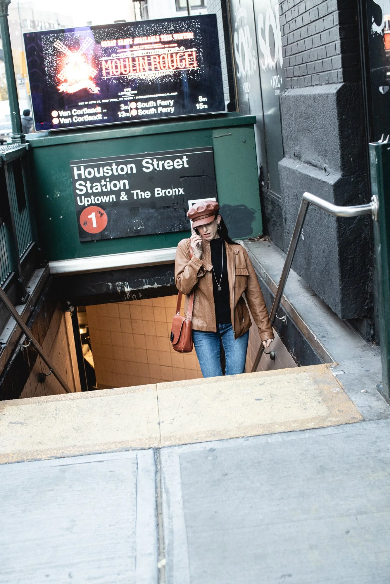 A woman with glasses, wearing a brown leather jacket, blue jeans, and a brown hat, walking up the stairs from Houston Street Station in New York City and talking on her cellphone.