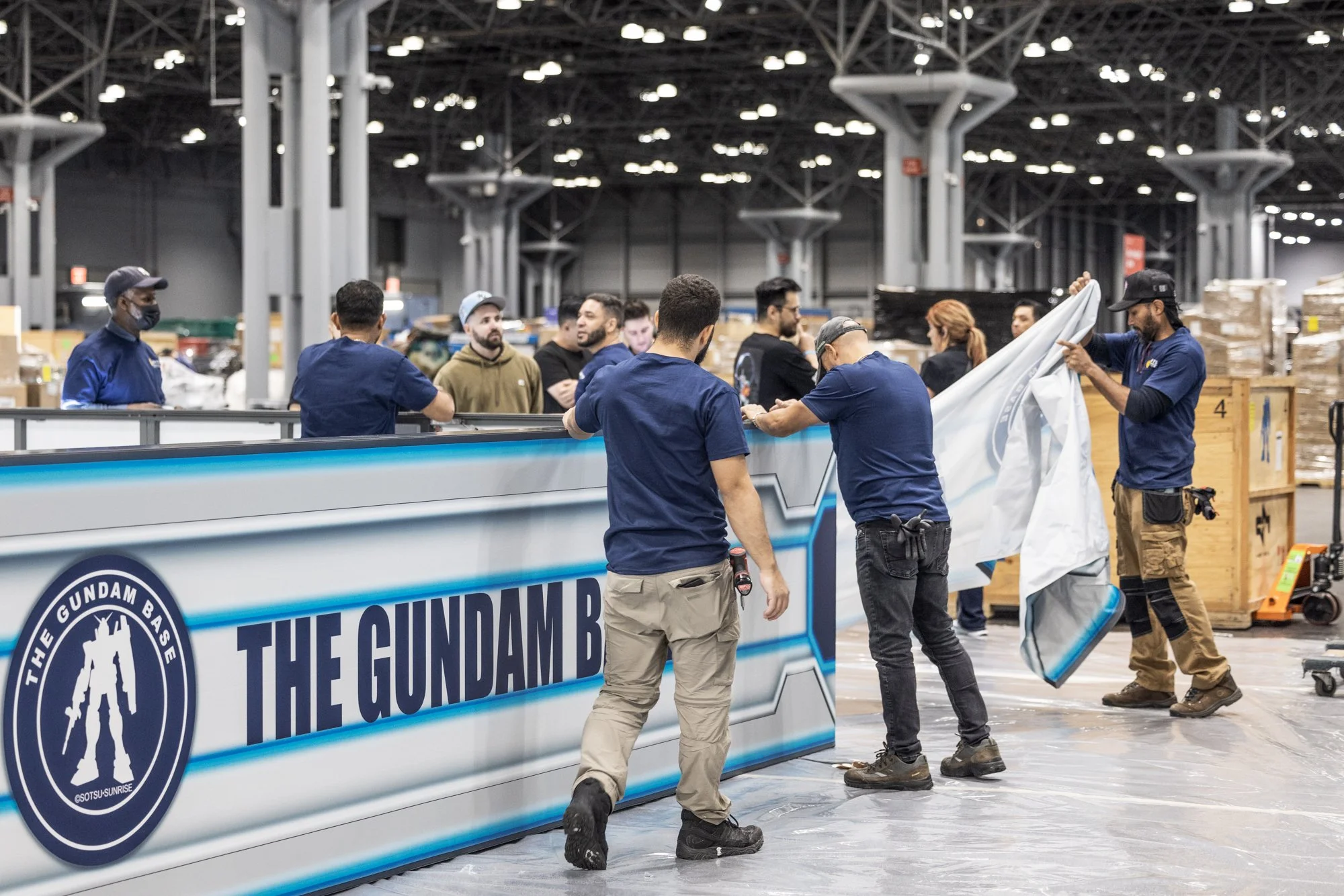 Crew members install a large Gundam Base display inside a convention center exhibition hall.