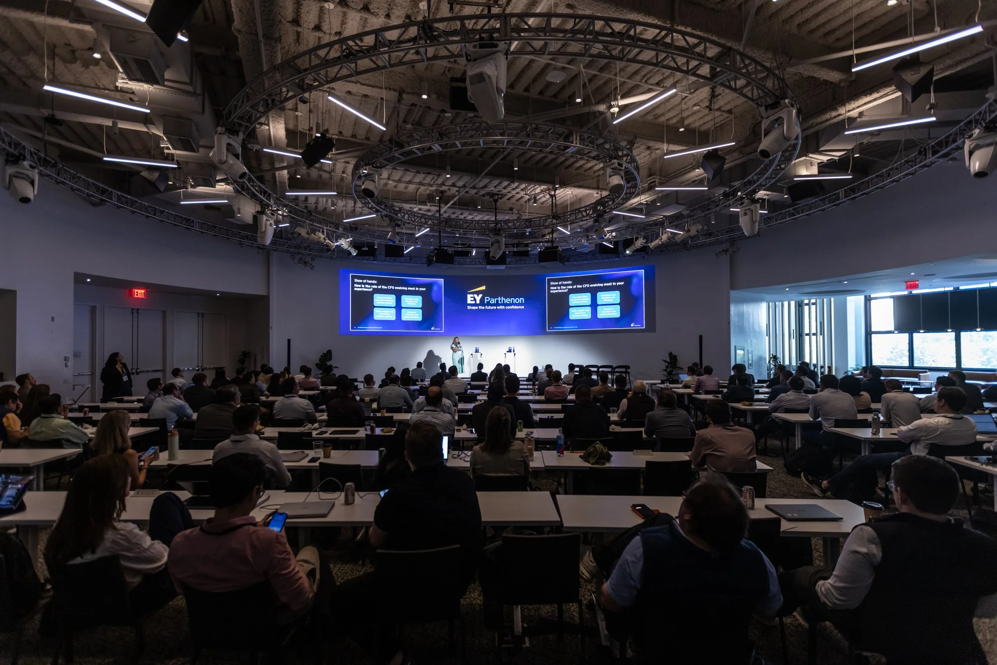 Large conference room filled with seated attendees and a presenter at the front. The room features high ceilings, a circular lighting and speaker rig, and two large screens displaying a presentation with the EY logo.