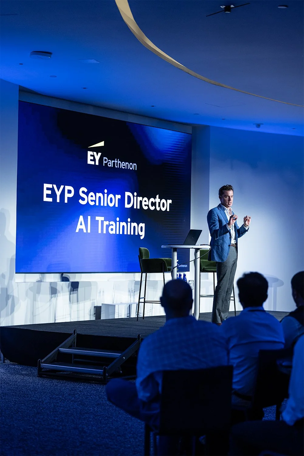 A man in a blue blazer giving a presentation on stage at a conference with a large screen behind him displaying "EY Parthenon, EYP Senior Director, AI Training." There are audience members seated facing the stage.