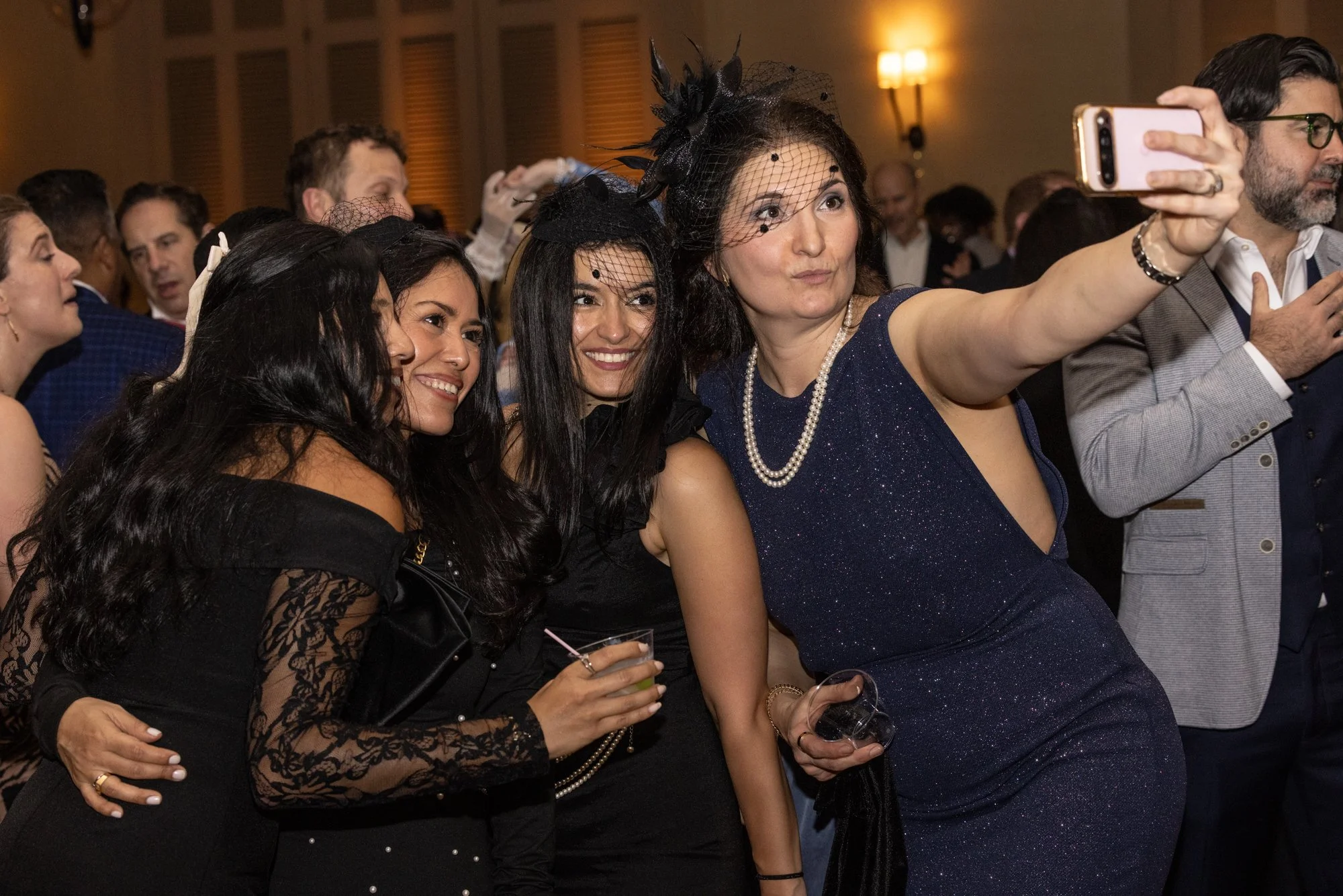 Group of women and men dressed in formal attire, taking a selfie at an event, with warm hotel lighting in the background.