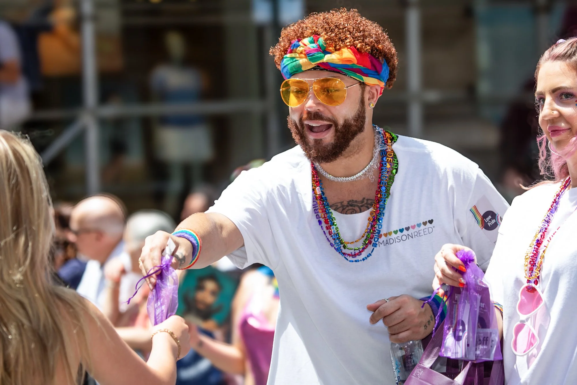 People celebrating at a Pride parade, including a bearded man wearing a rainbow headband, yellow sunglasses, and colorful necklaces.