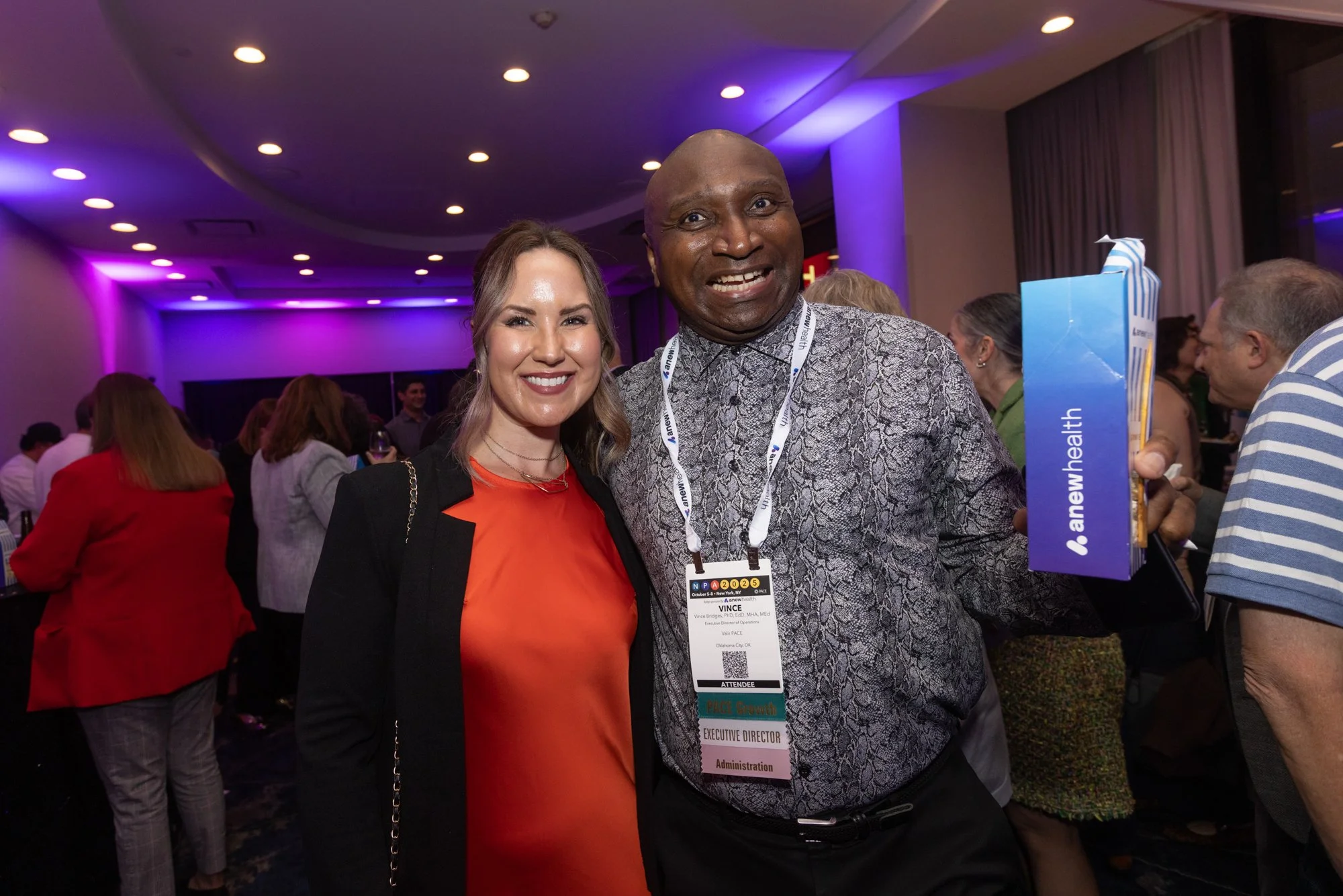 Two people smiling at a conference or event. The woman is wearing an orange dress with a black blazer, and the man is wearing a patterned shirt. Both have event badges hanging around their necks. There are other attendees in the background, and purpl