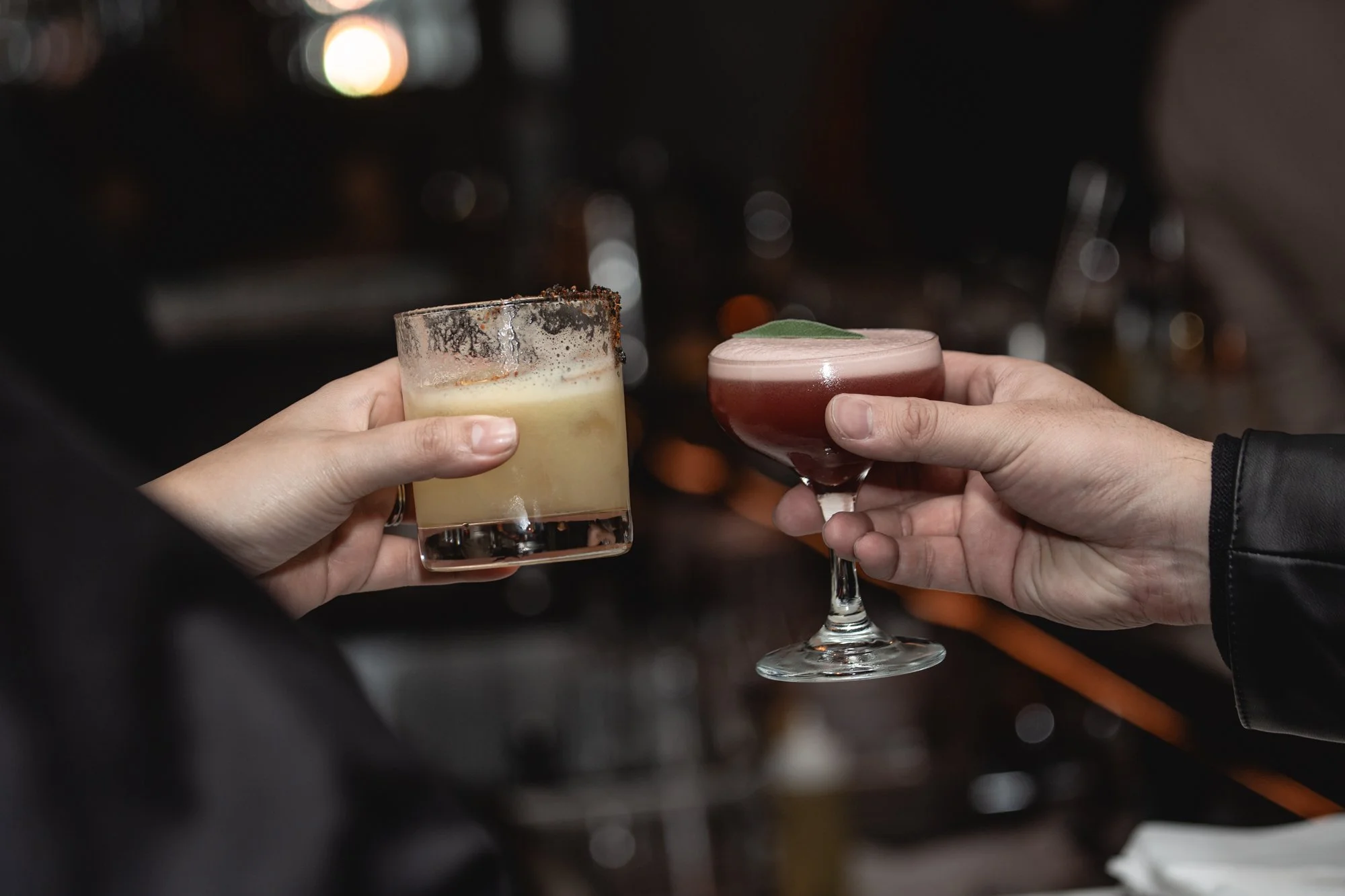 Two people holding cocktails in a toast at a bar, one with a frothy pink drink with a leaf garnish and the other with a yellowish drink with a salted rim and possibly a spice topping.