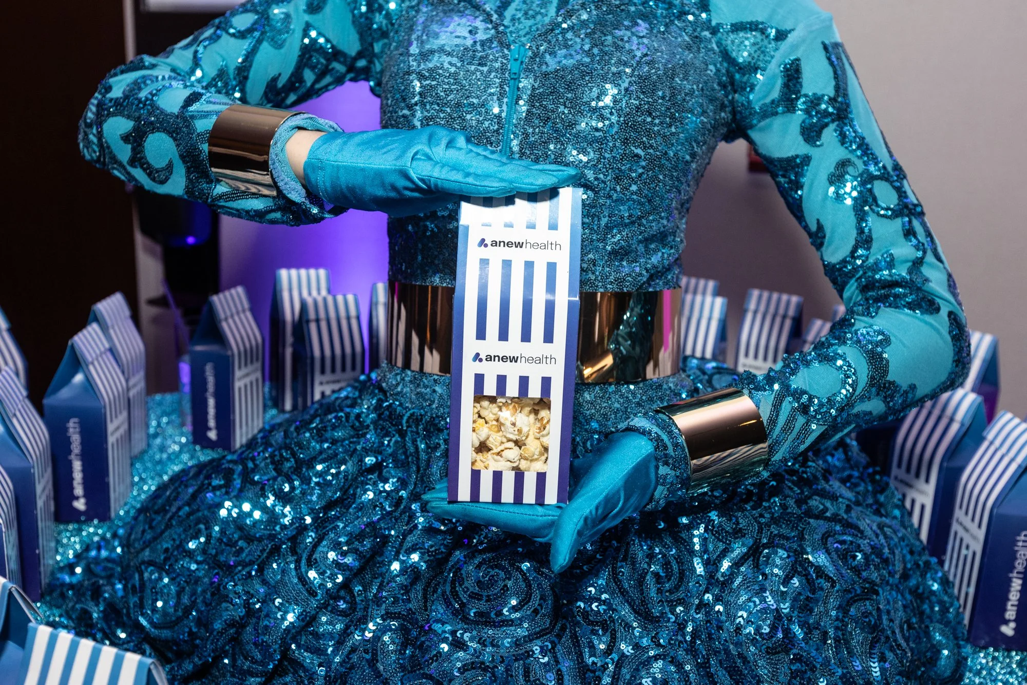A person dressed in a shiny, sequined blue costume with long sleeves, a high collar, and metallic bands on their wrists holding a striped box of popcorn. They are also wearing a matching blue glove on one hand. The box has a logo that says 'anew heal
