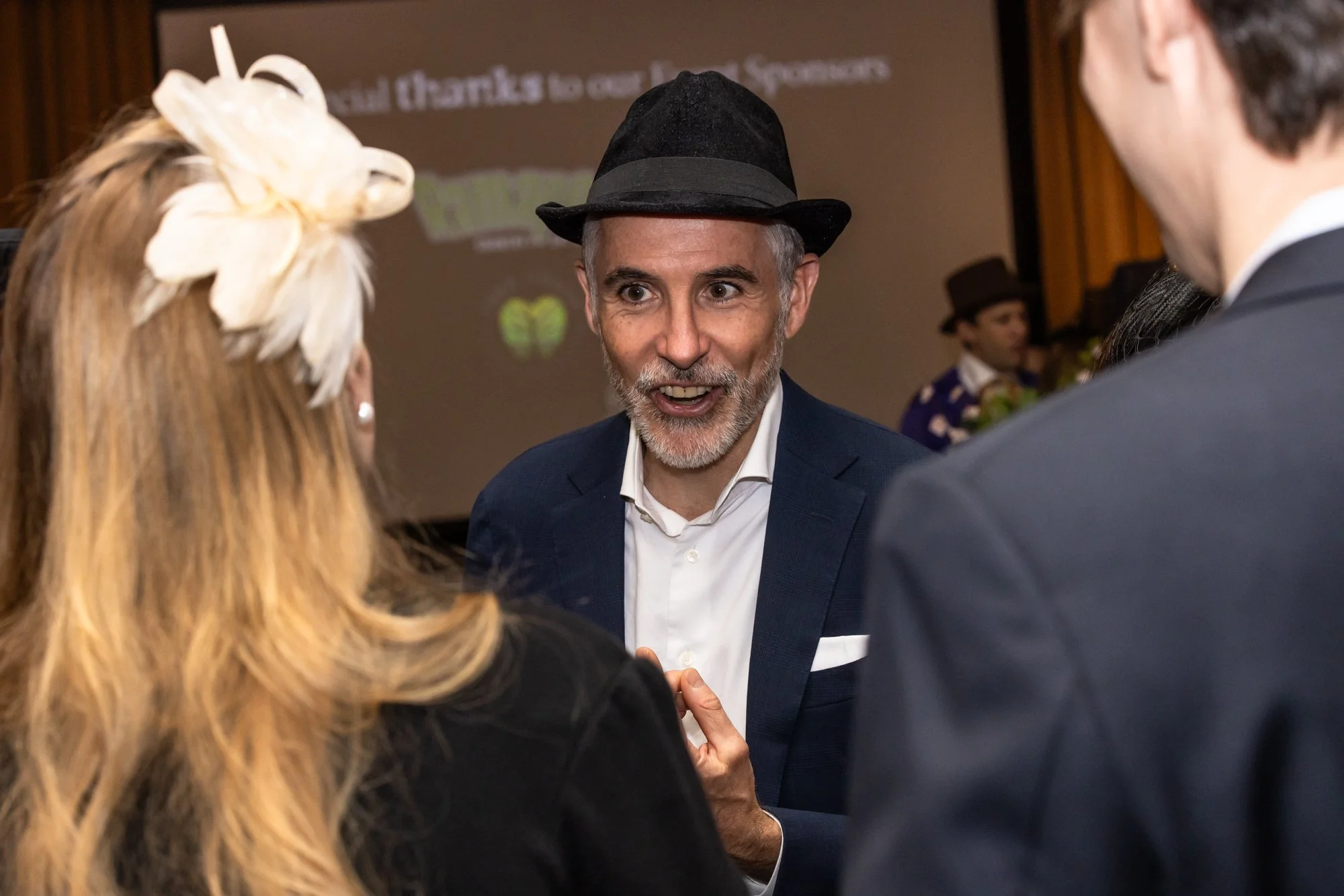 A man in a dark suit and hat talking to a woman with blonde hair wearing a flower in her hair and a dark outfit at a social event.