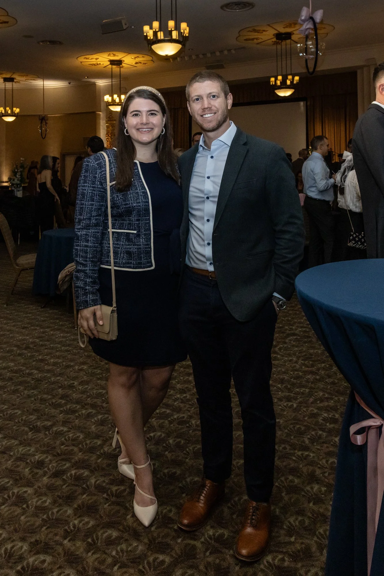 A woman and a man dressed in formal attire, standing close together and smiling at an indoor event with warm lighting and several other guests in the background.