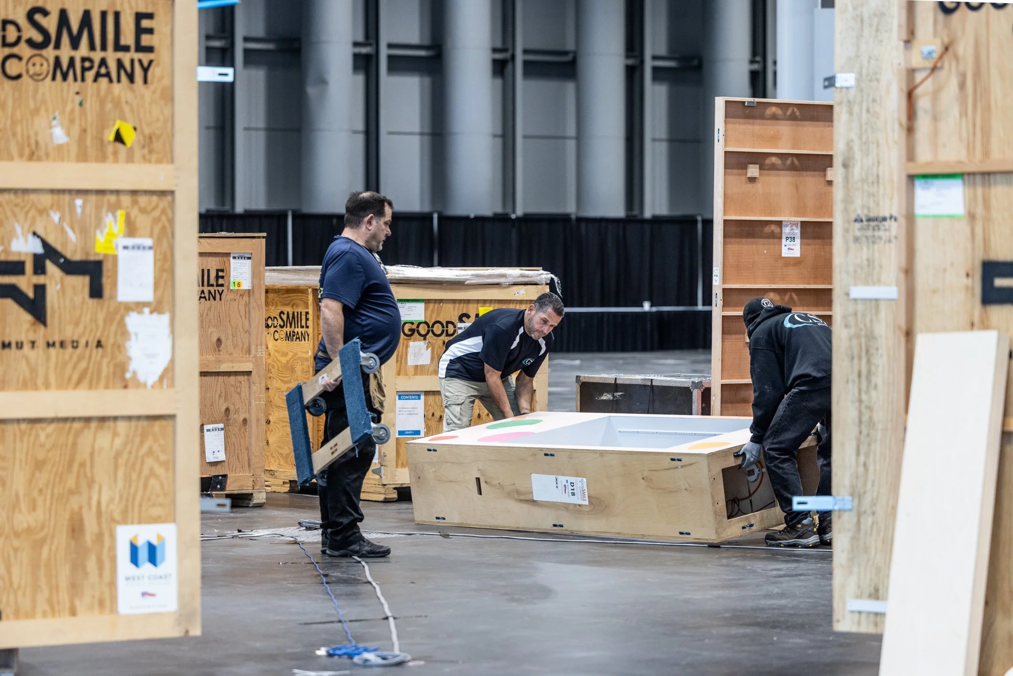 Three workers move a large wooden crate through an indoor warehouse surrounded by shipping cases.