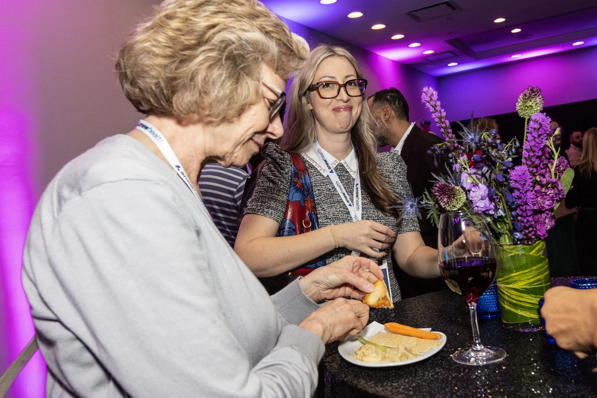 Two women are sitting at a table at an event with purple lighting. One woman with short, light-colored hair is eating a piece of bread, and the other woman with long, gray hair and glasses is holding a glass of red wine. The table has a floral arrang