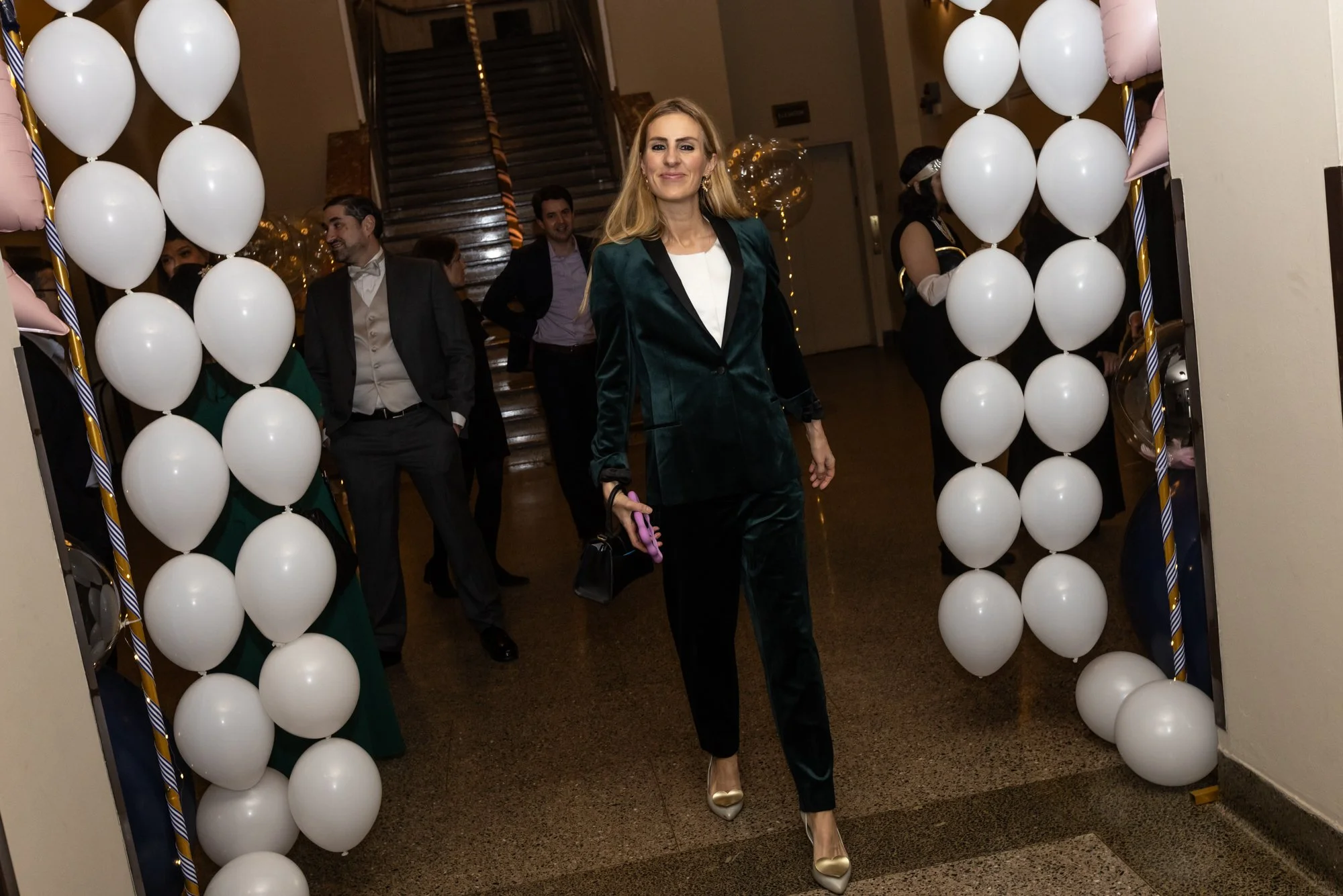 A woman with blond hair wearing a dark green velvety suit and gold heels walking through a balloon arch at an indoor event. Several people, mostly men in suits, are in the background near a staircase.
