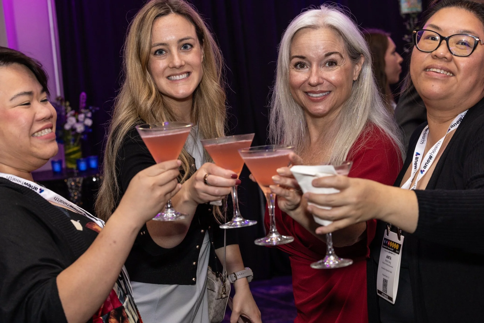 Group of women at an event raising pink cocktails in a toast, smiling and celebrating together, wearing event lanyards.
