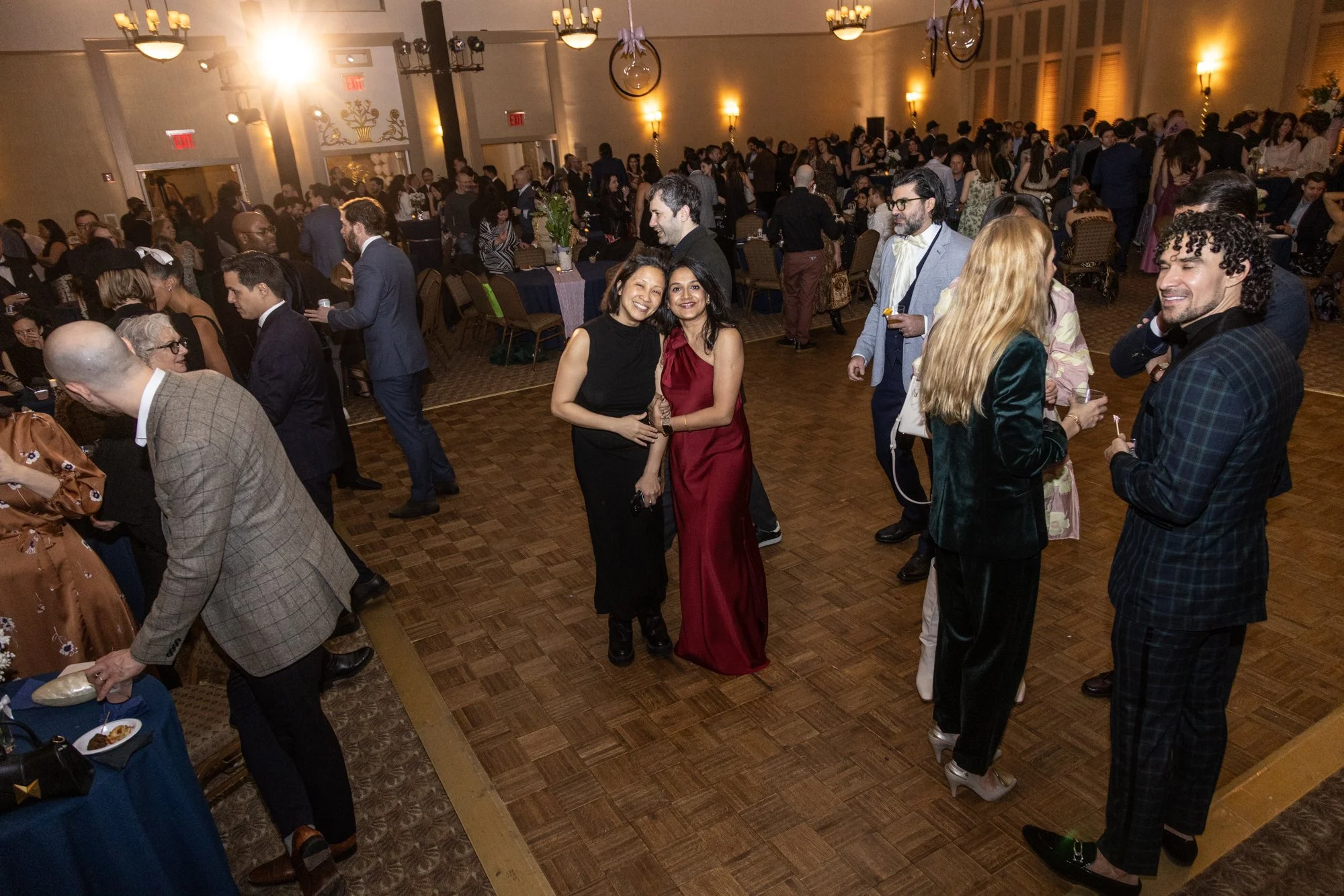 People socializing at a large indoor event with round tables and a dance floor, decorated in elegant style.
