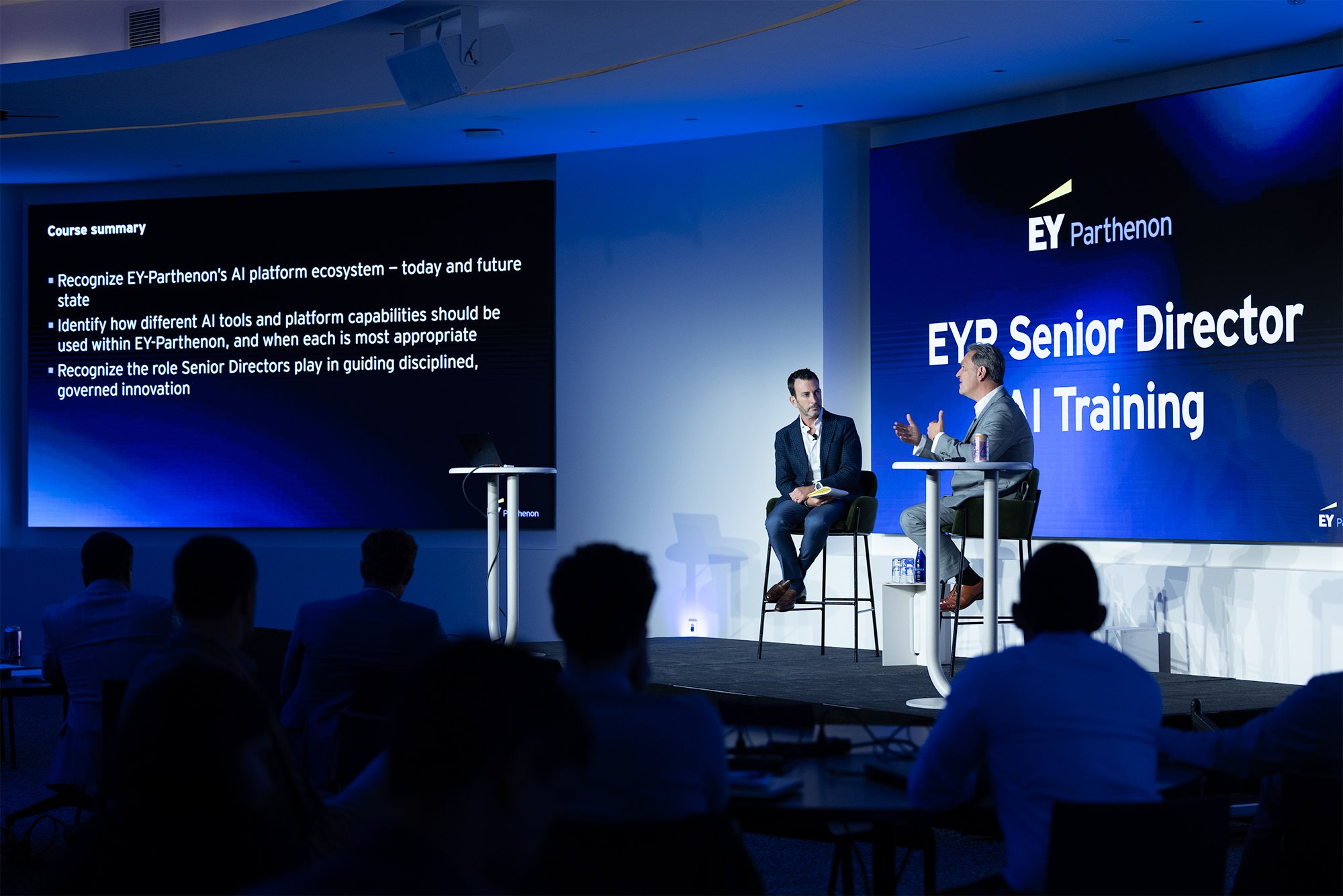 A conference room with a stage where two men are seated and speaking during a presentation. Large screens display the EY Parthenon logo and presentation slides related to AI platform ecosystem and senior director roles. Audience members are seated in