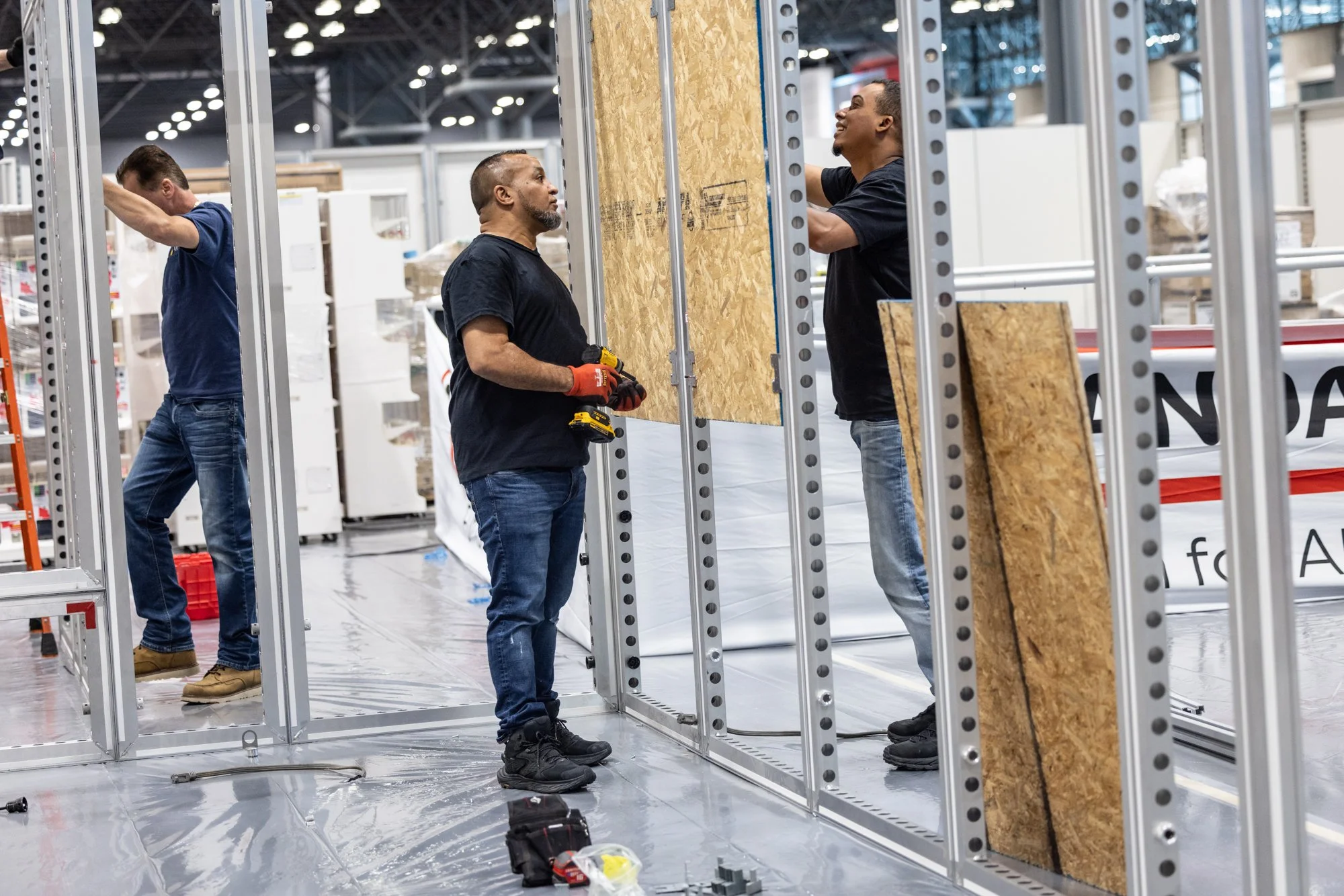 Three workers assemble a metal-framed trade show booth with plywood panels in an exhibition hall.
