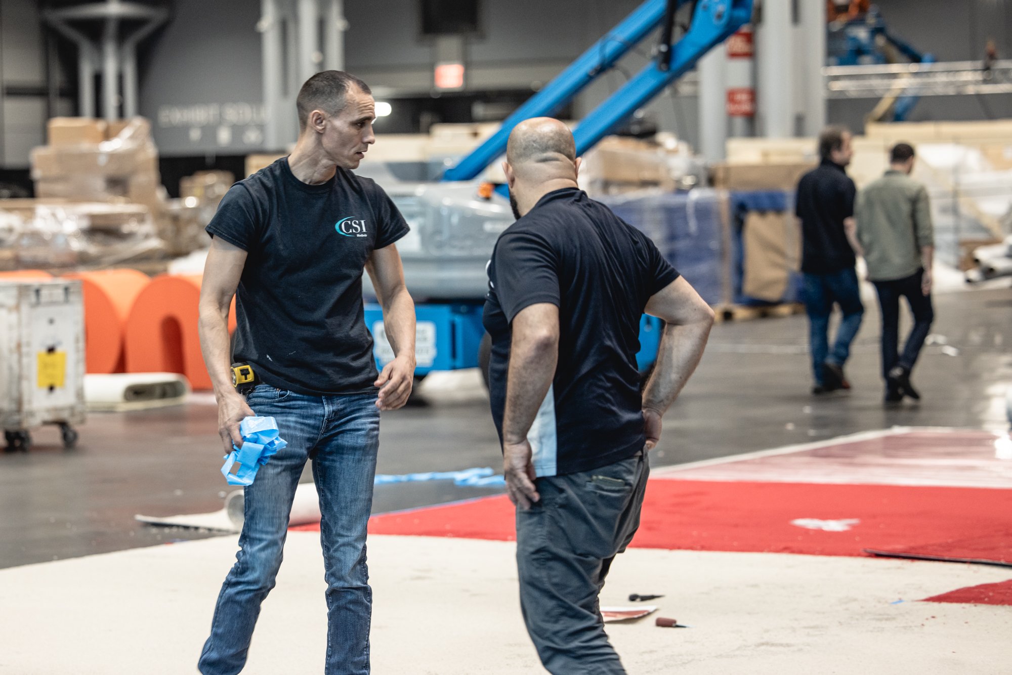 Two workers wearing black shirts talk inside a warehouse with blue lifting equipment and stacked materials.