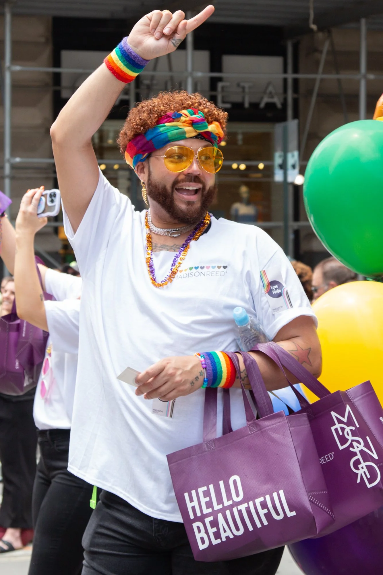 Person wearing rainbow accessories and sunglasses raises a hand at a Pride event while carrying purple shopping bags beside colorful balloons.