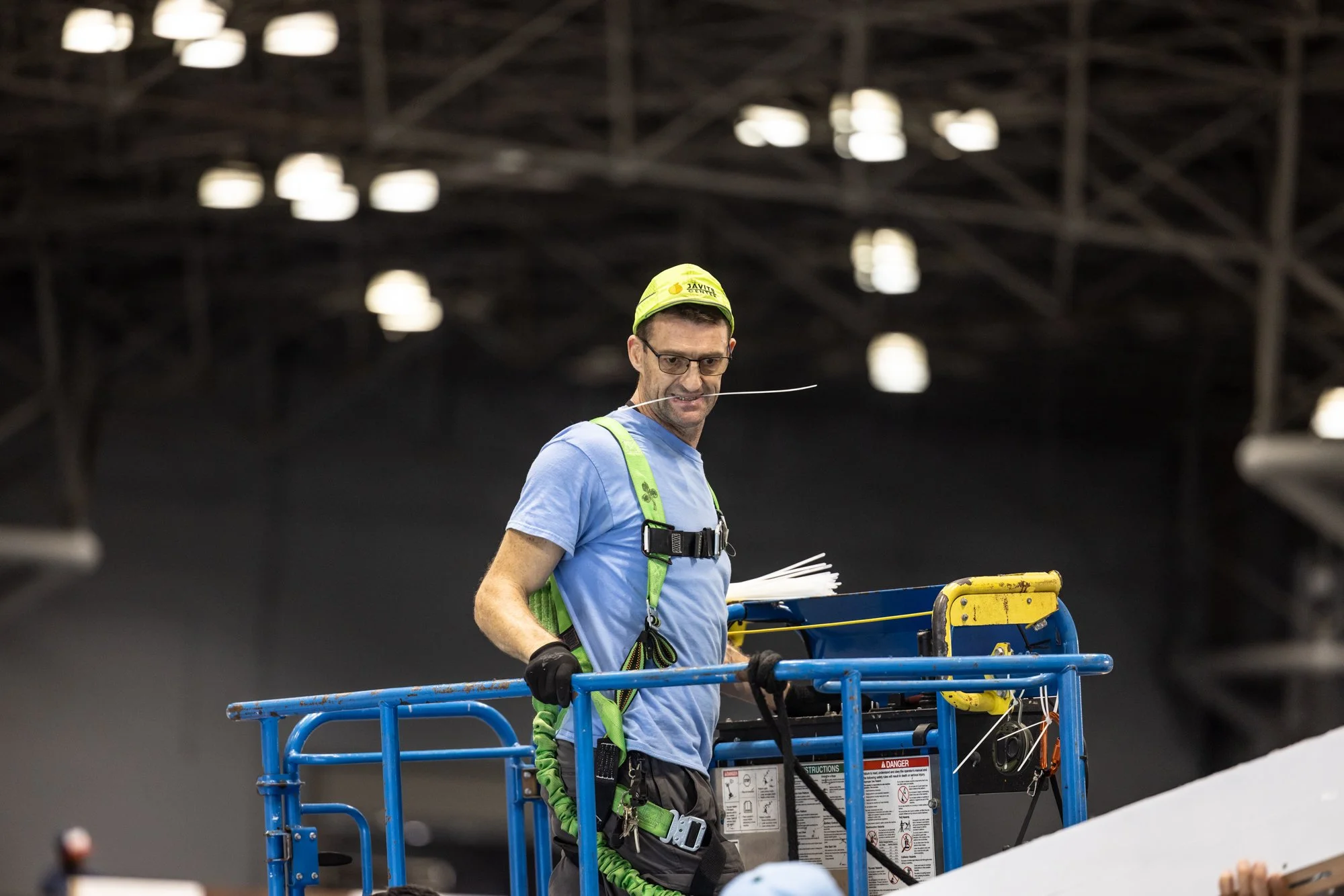 Worker wearing safety harness stands in a blue lift beneath stadium lights.