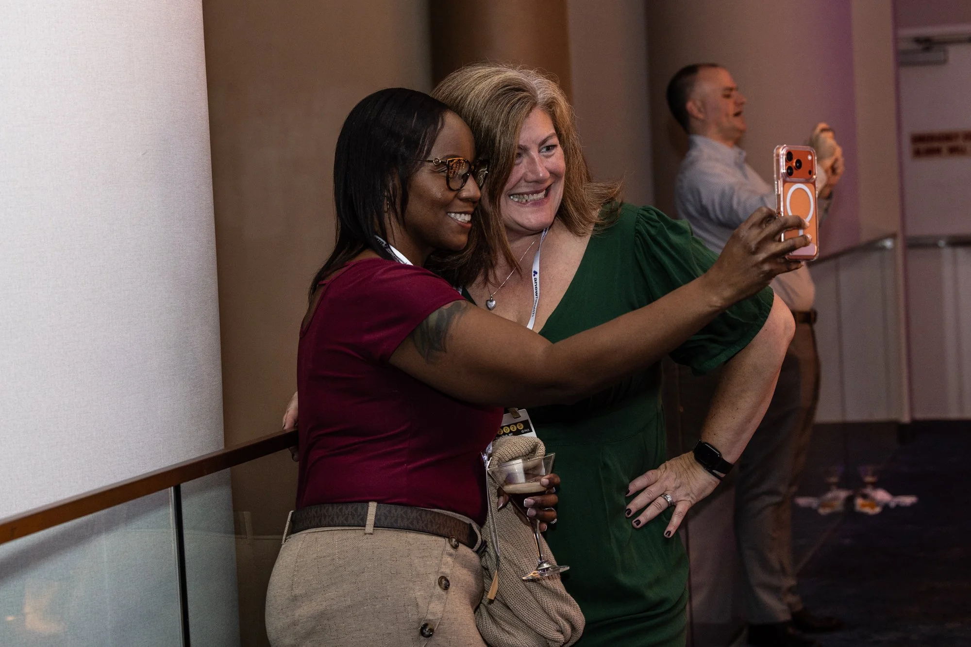 Two women taking a selfie at an event, smiling and standing close together, with other attendees in the background.