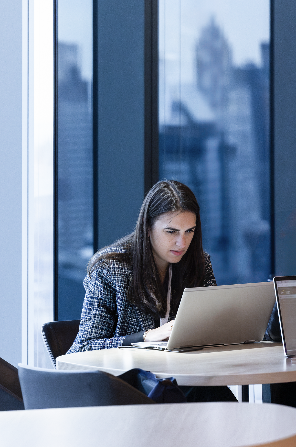 A woman with long dark hair working on a laptop at a conference room table, with large window reflections behind her.