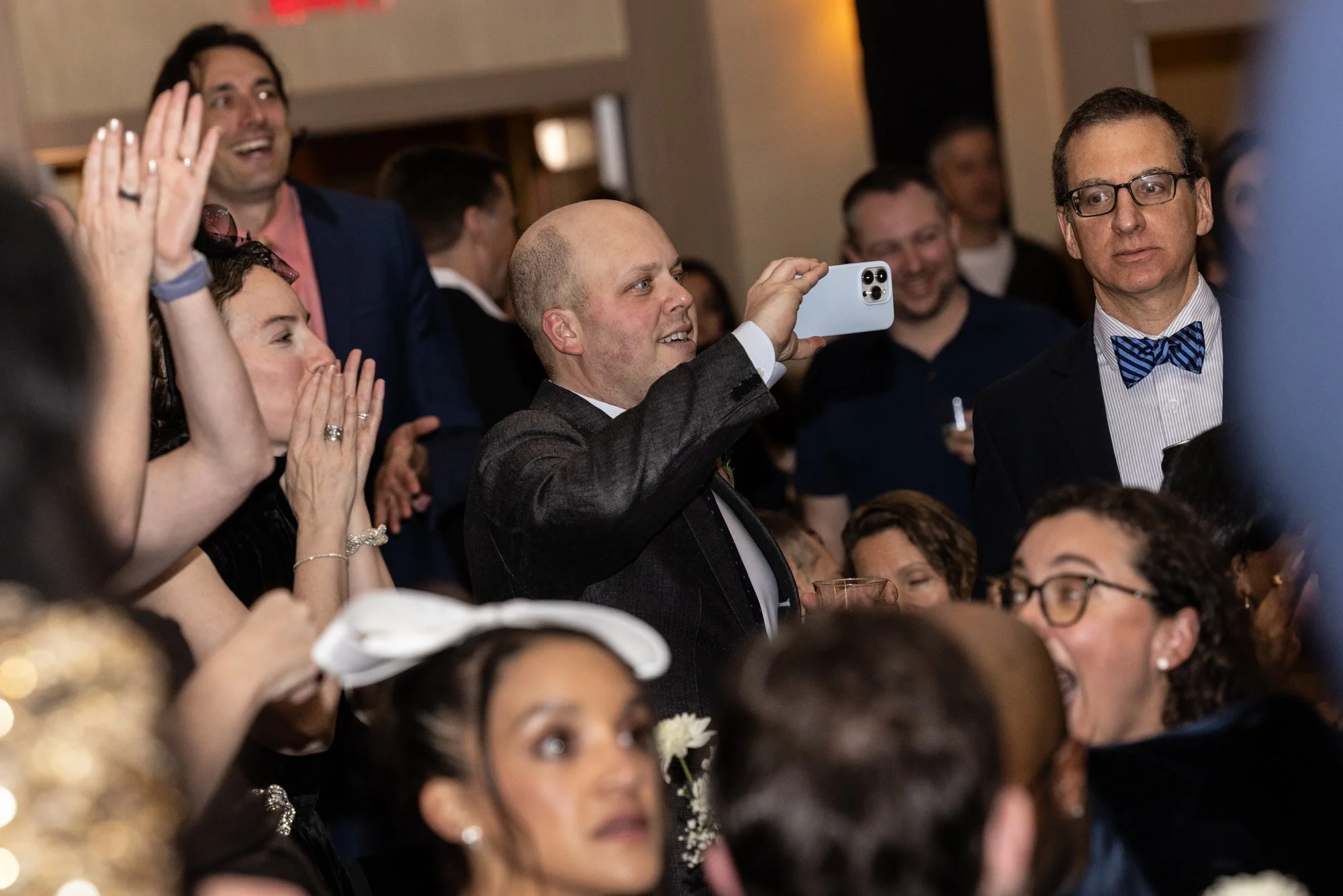 A group of people at a formal event, some clapping and smiling, with one man taking a photo with his phone. The setting appears to be a banquet or celebration.