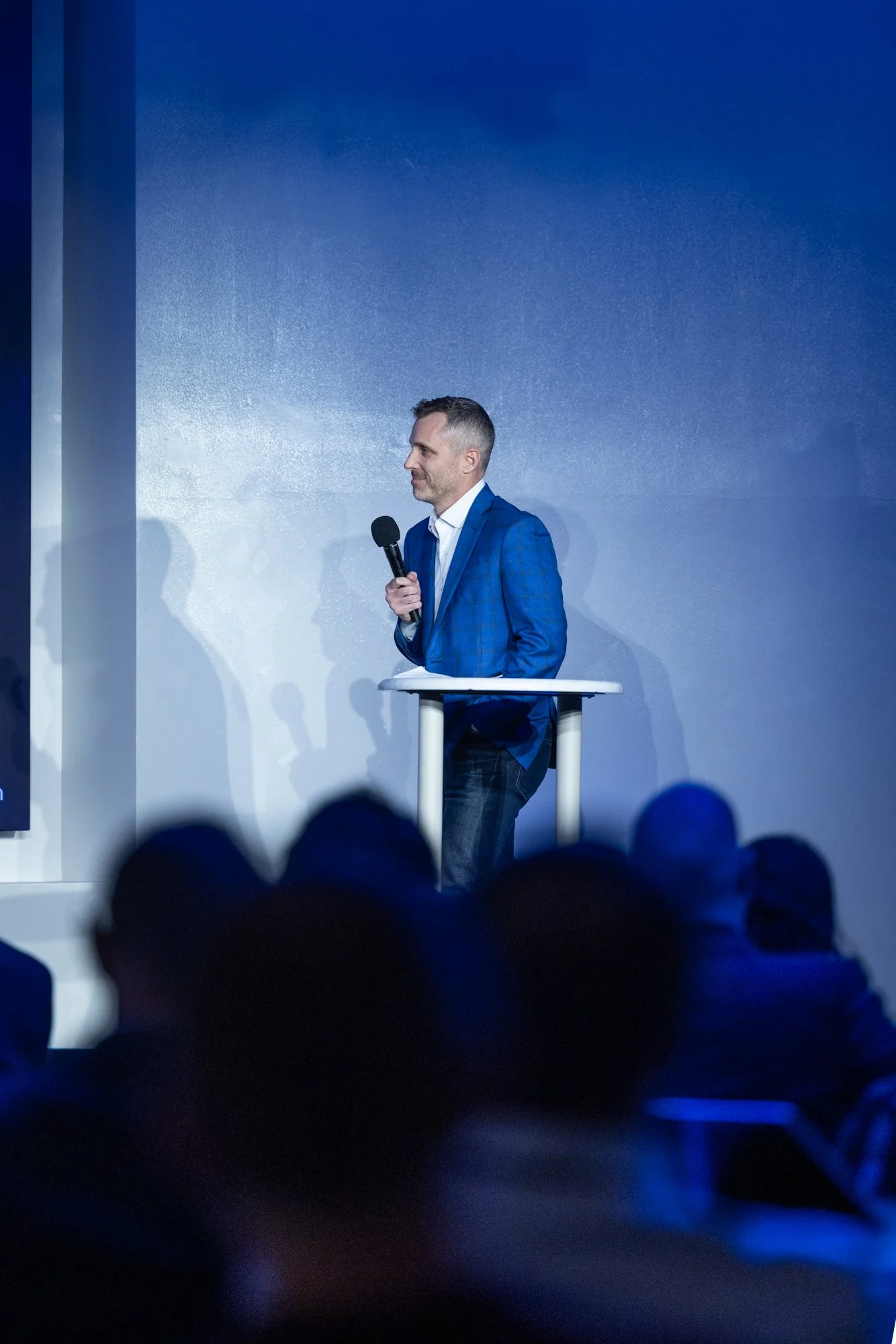A man in a blue suit standing at a podium with a microphone during a presentation or speech, with a blue background and audience visible in the foreground.