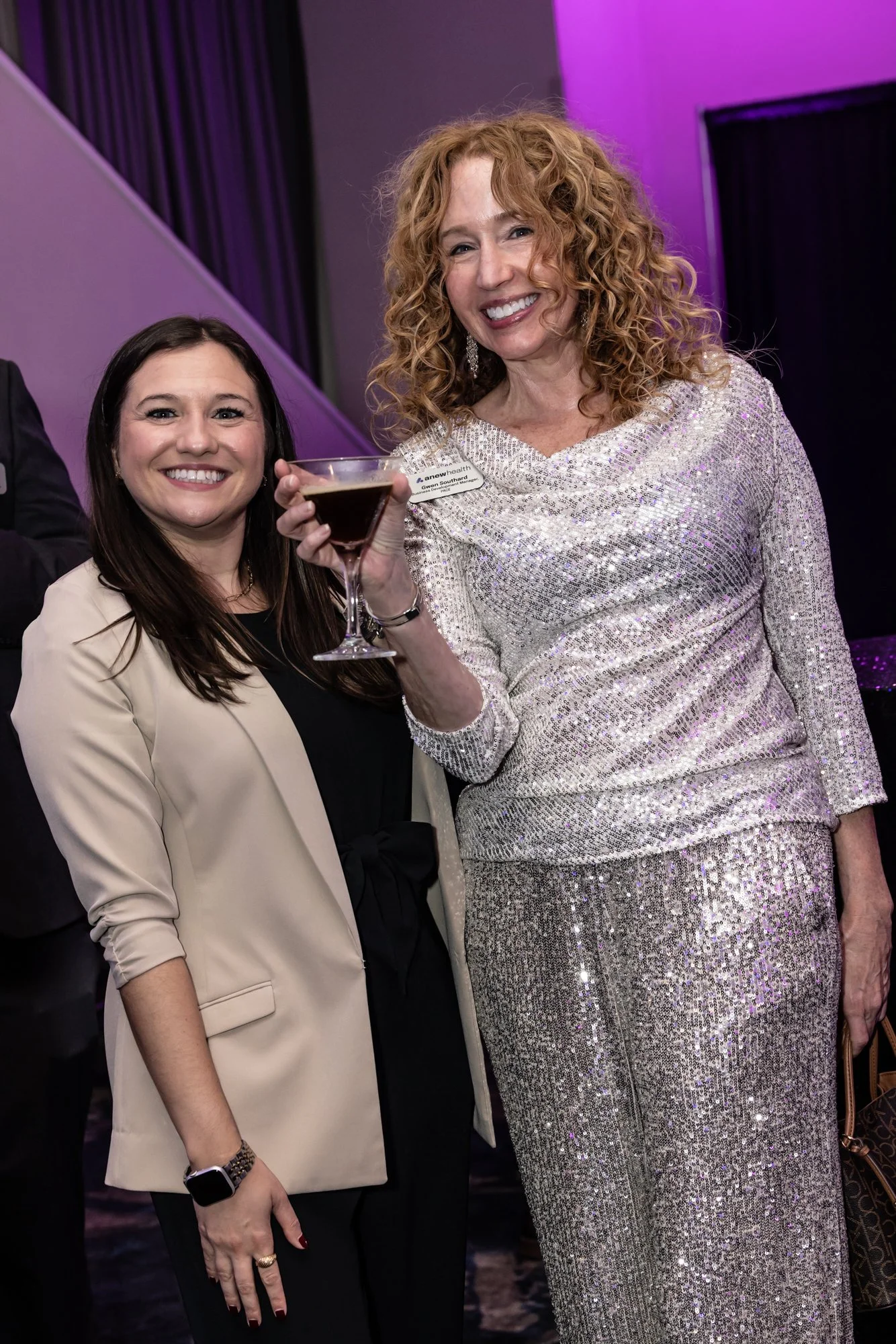 Two women smiling at an event, one holding a martini glass, with purple lighting in the background.