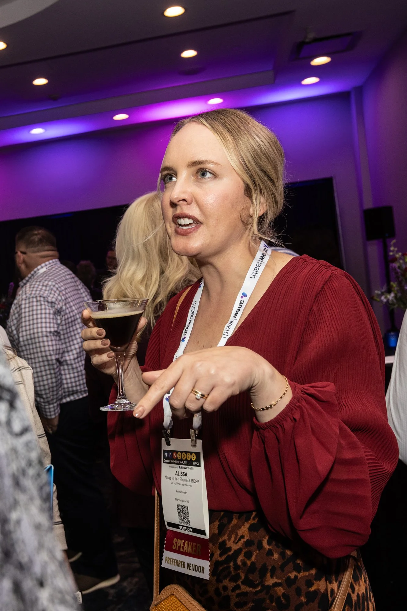Woman wearing a red top and lanyard holding a cocktail glass at a professional conference or event, with other attendees visible in the background.