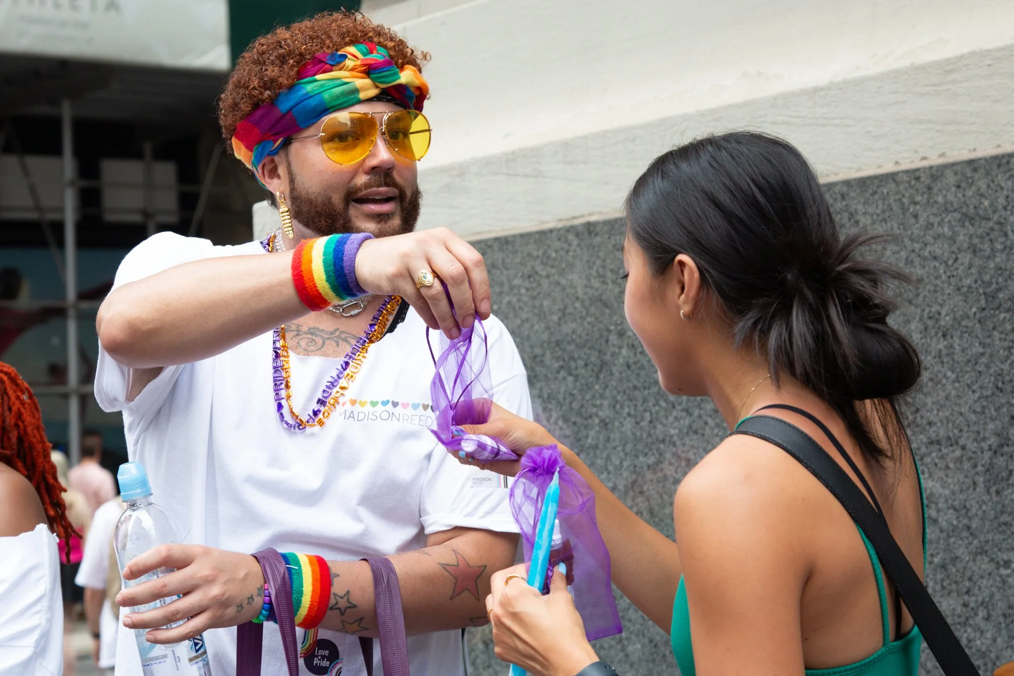 Person wearing rainbow accessories and sunglasses handing a purple ribbon to a woman at a Pride event