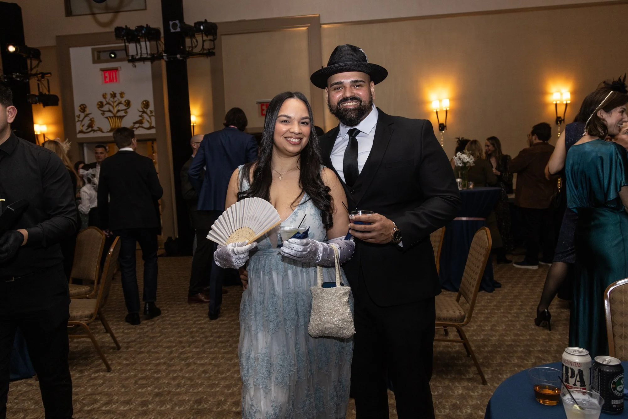 A smiling couple at a formal event, the woman in a blue dress holding a hand fan and a small purse, the man in a black suit with a tie and a wide-brimmed black hat, standing in a decorated banquet hall with other guests in the background.