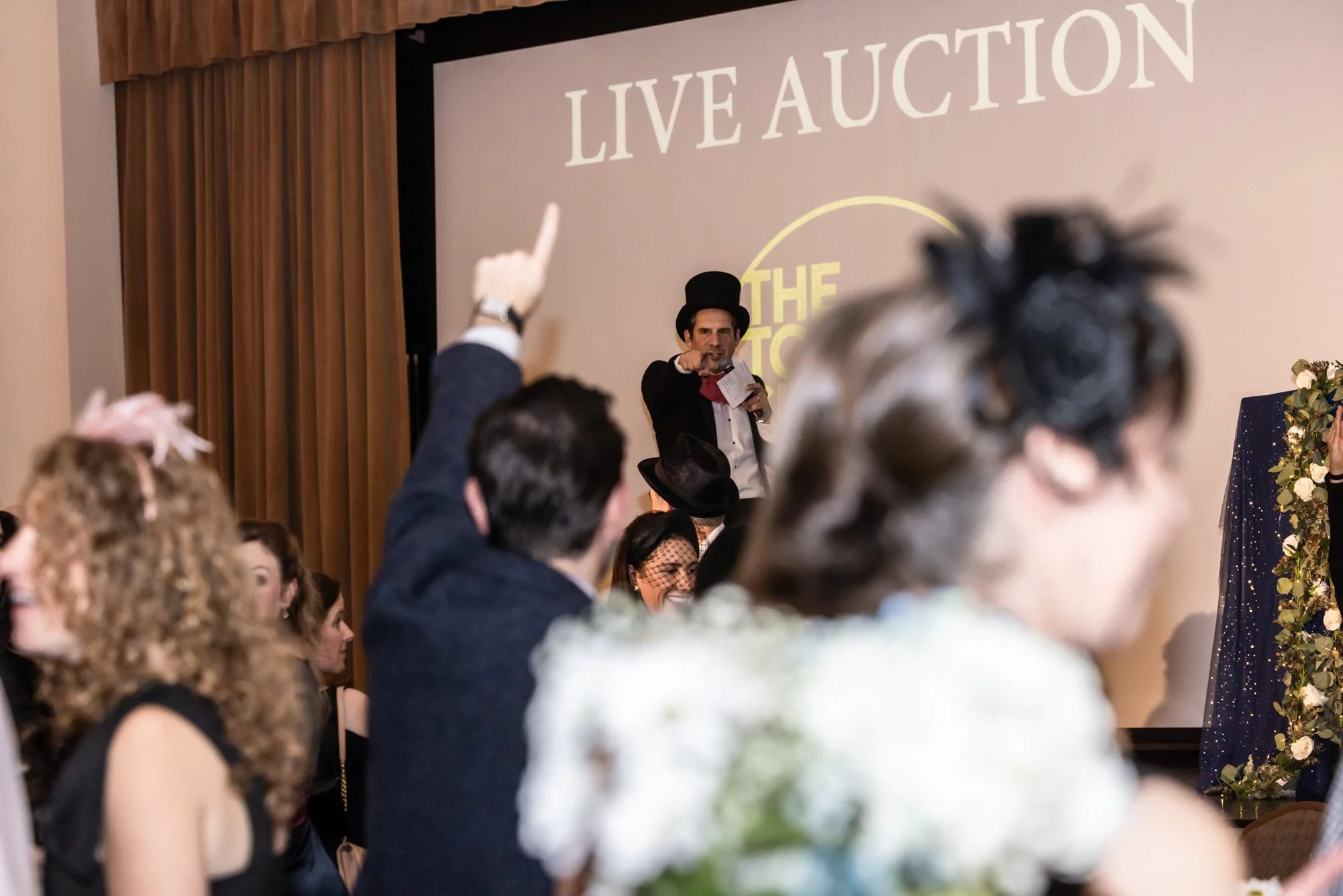 A man dressed as a ringmaster, wearing a black top hat and a tuxedo, speaking at a live auction event. He is gesturing towards the audience who are attentively listening. The background shows a large screen with the words 'LIVE AUCTION', and there ar
