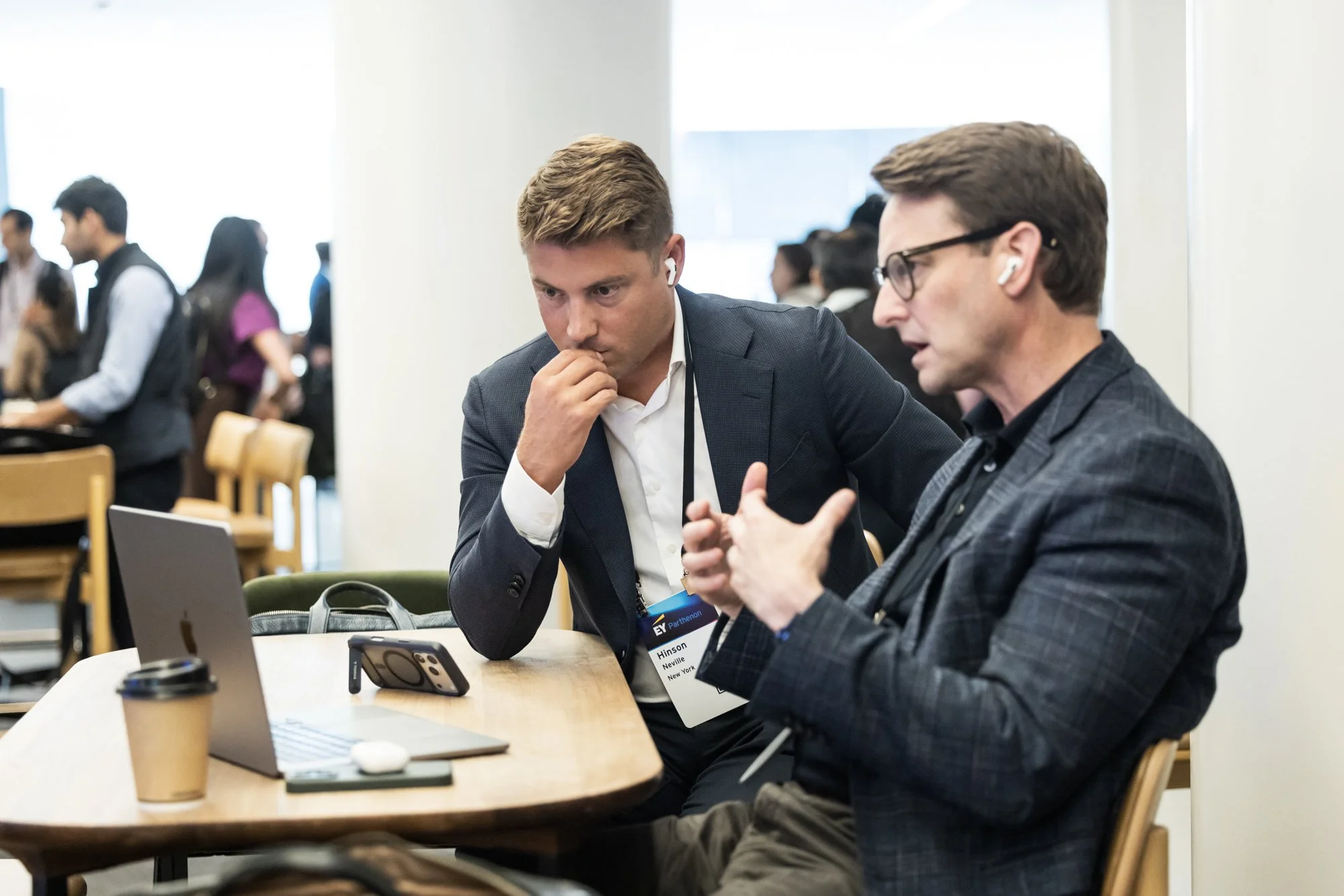 Two men in suits sitting at a table using wireless earbuds, engaged in conversation, in a busy conference or networking event.