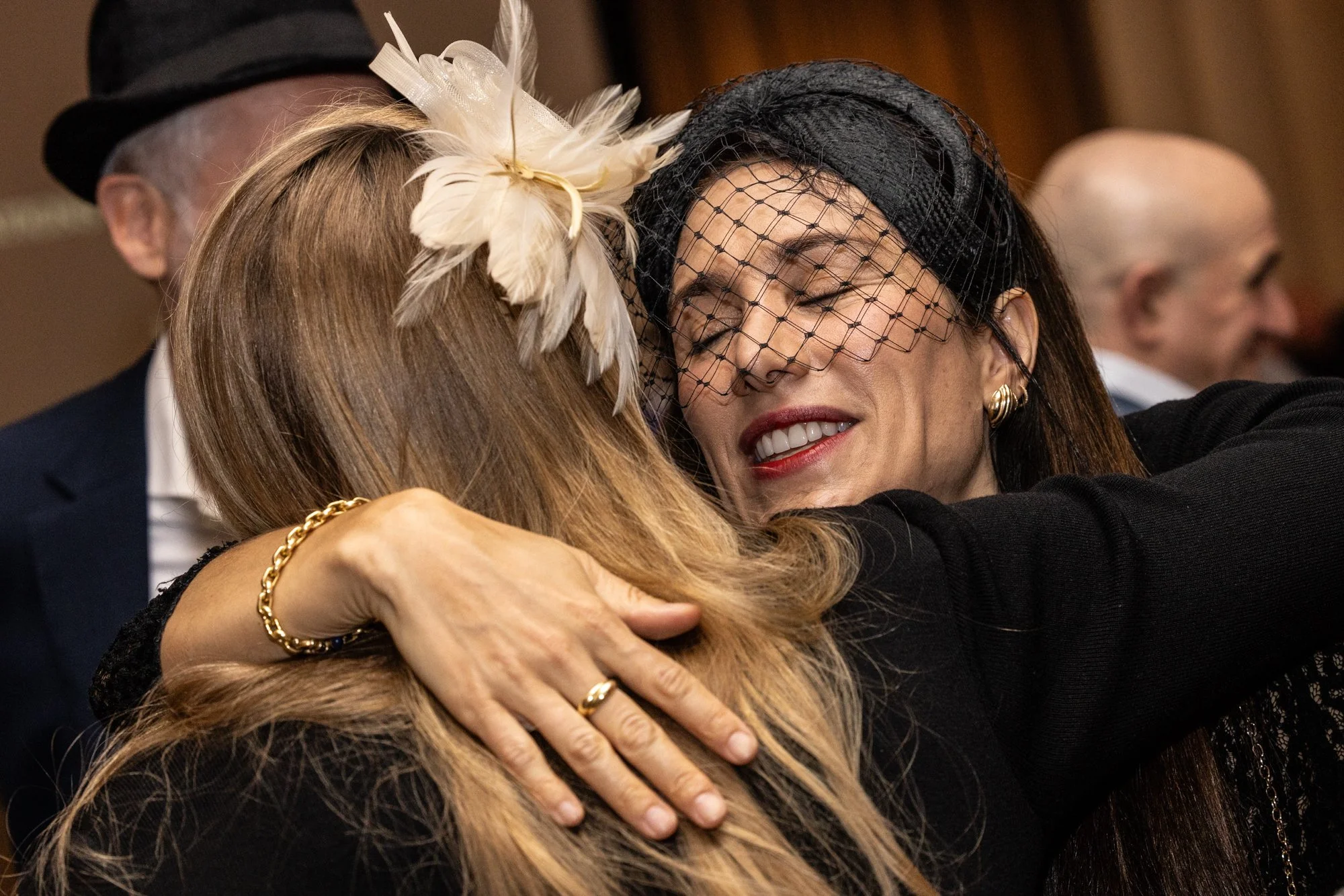 People hugging at a social gathering, with some wearing fancy attire and hats, including a woman wearing a black netted fascinator.