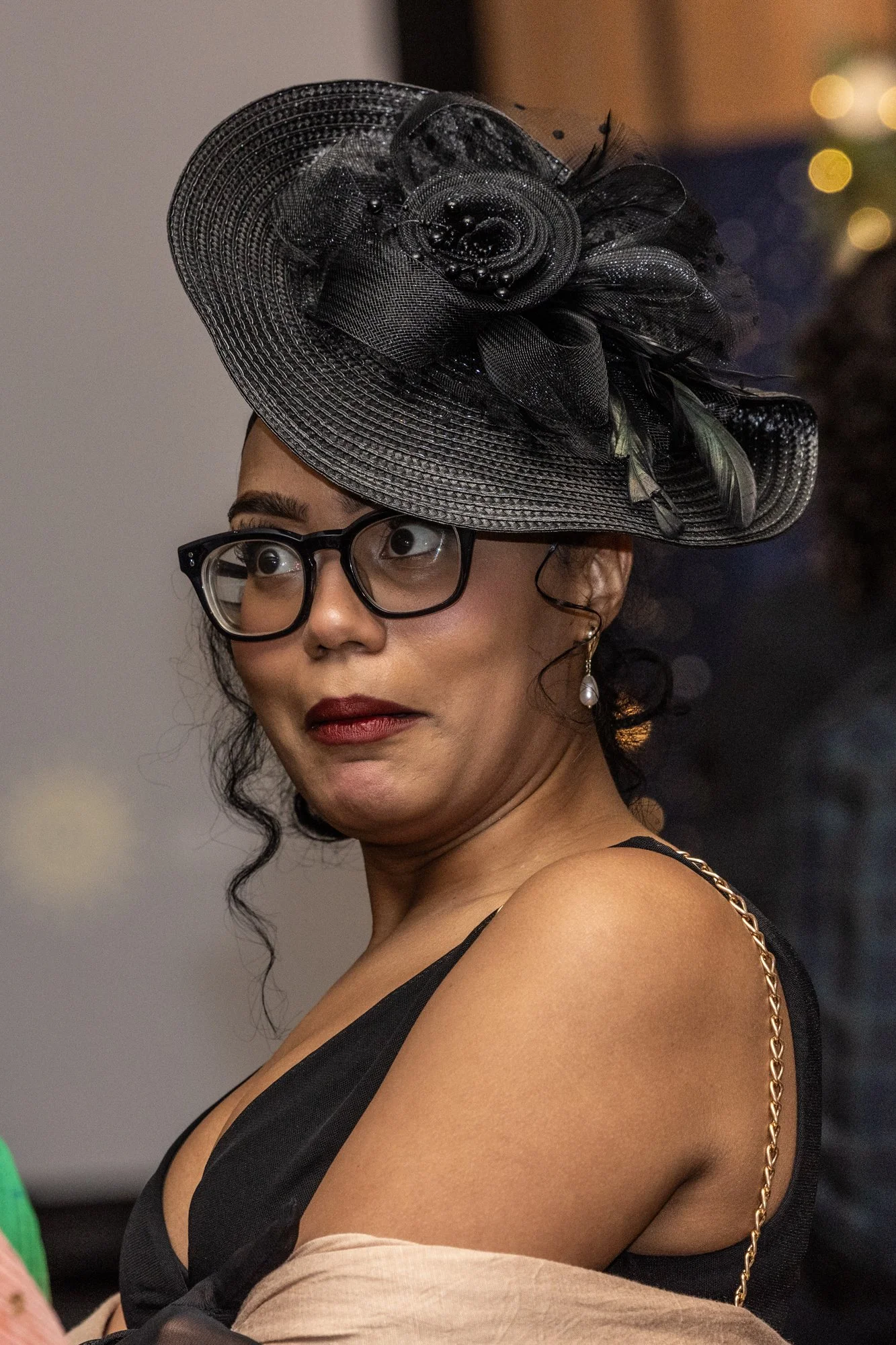 A woman with dark hair, wearing black glasses, a large black hat with decorative feathers and netting, earrings, and a black dress, looking to her left with a neutral expression.