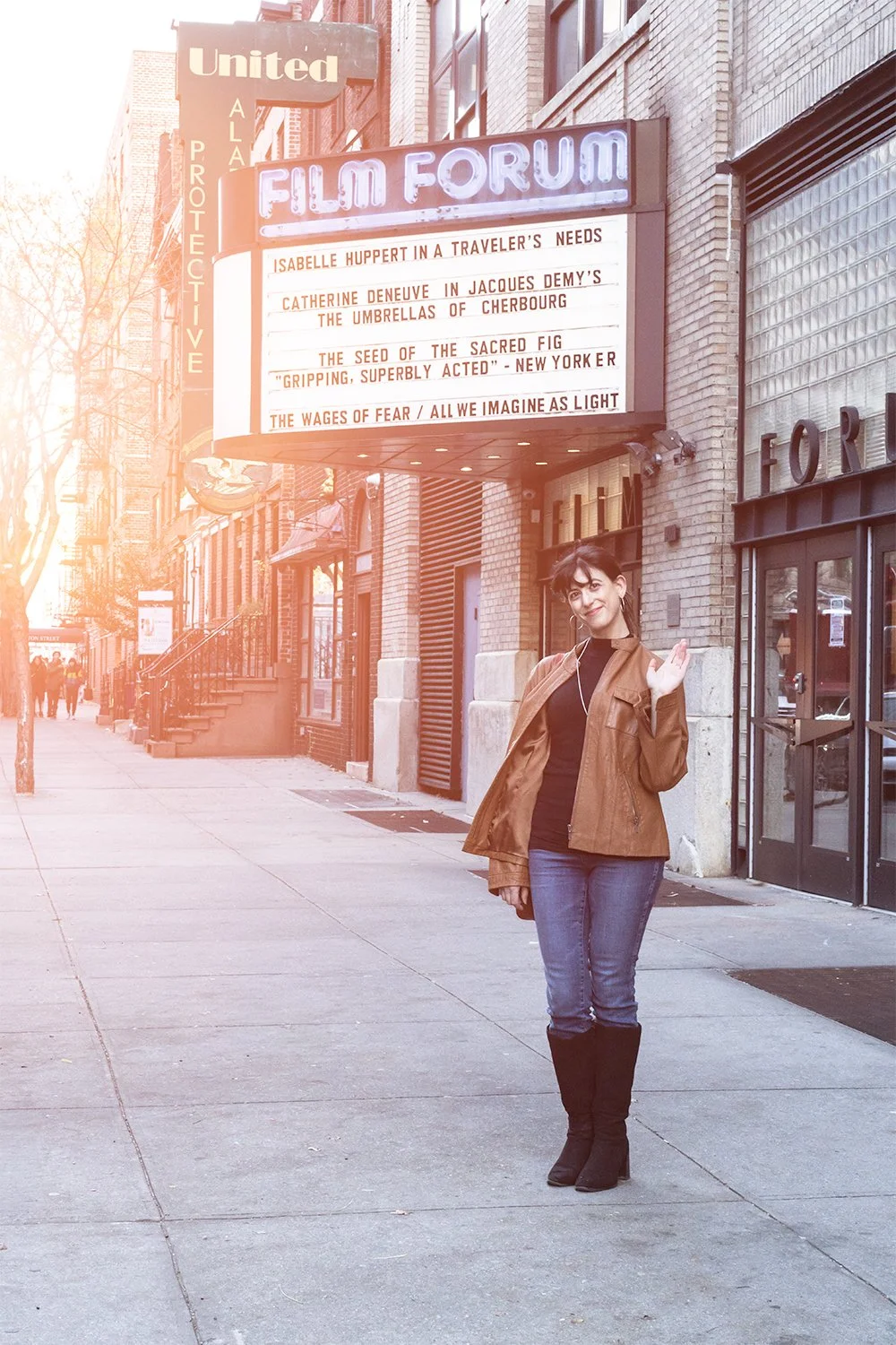 A woman with dark hair, wearing a brown jacket, black top, blue jeans, and black boots, waving and smiling on a city sidewalk in front of a marquee that reads 'Film Forum' with a list of film titles and reviews.