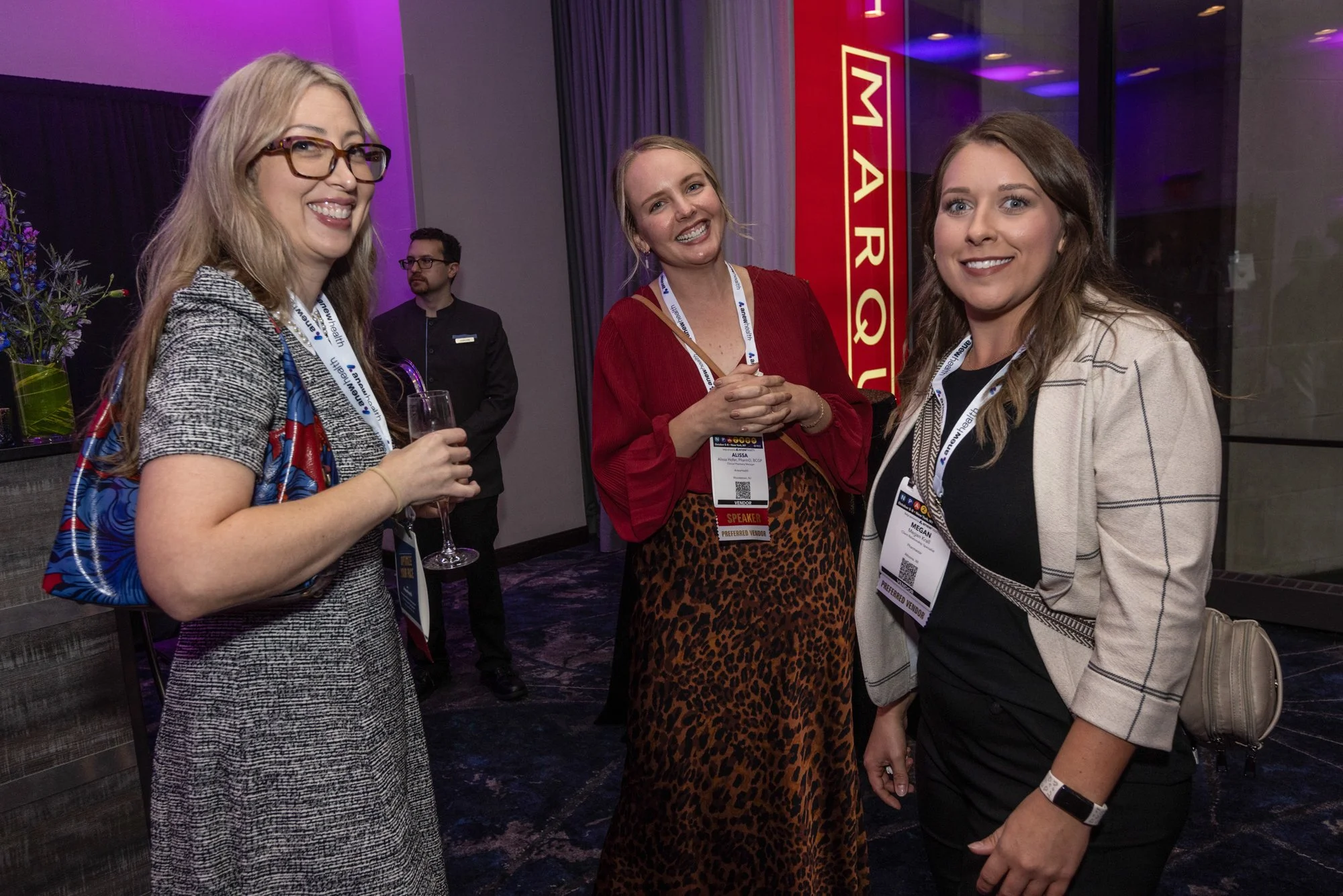 Three women smiling at a social event, wearing badges, indoors with purple lighting and a red sign in the background.
