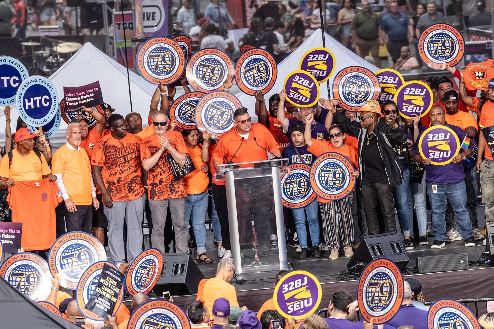 A crowd of protesters at a rally, many wearing orange shirts and holding signs with labor union logos, including SEIU and 32BJ, in an outdoor city setting with buildings and a stage in the background.