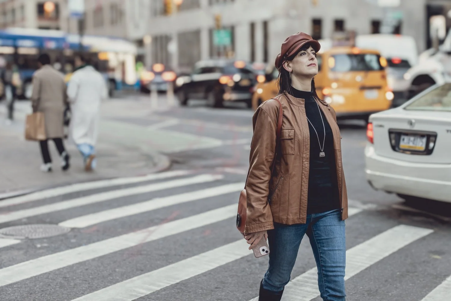 A woman crossing a busy crosswalk in an urban city, holding a smartphone, wearing a brown jacket, jeans, and a hat, with traffic and other pedestrians in the background.
