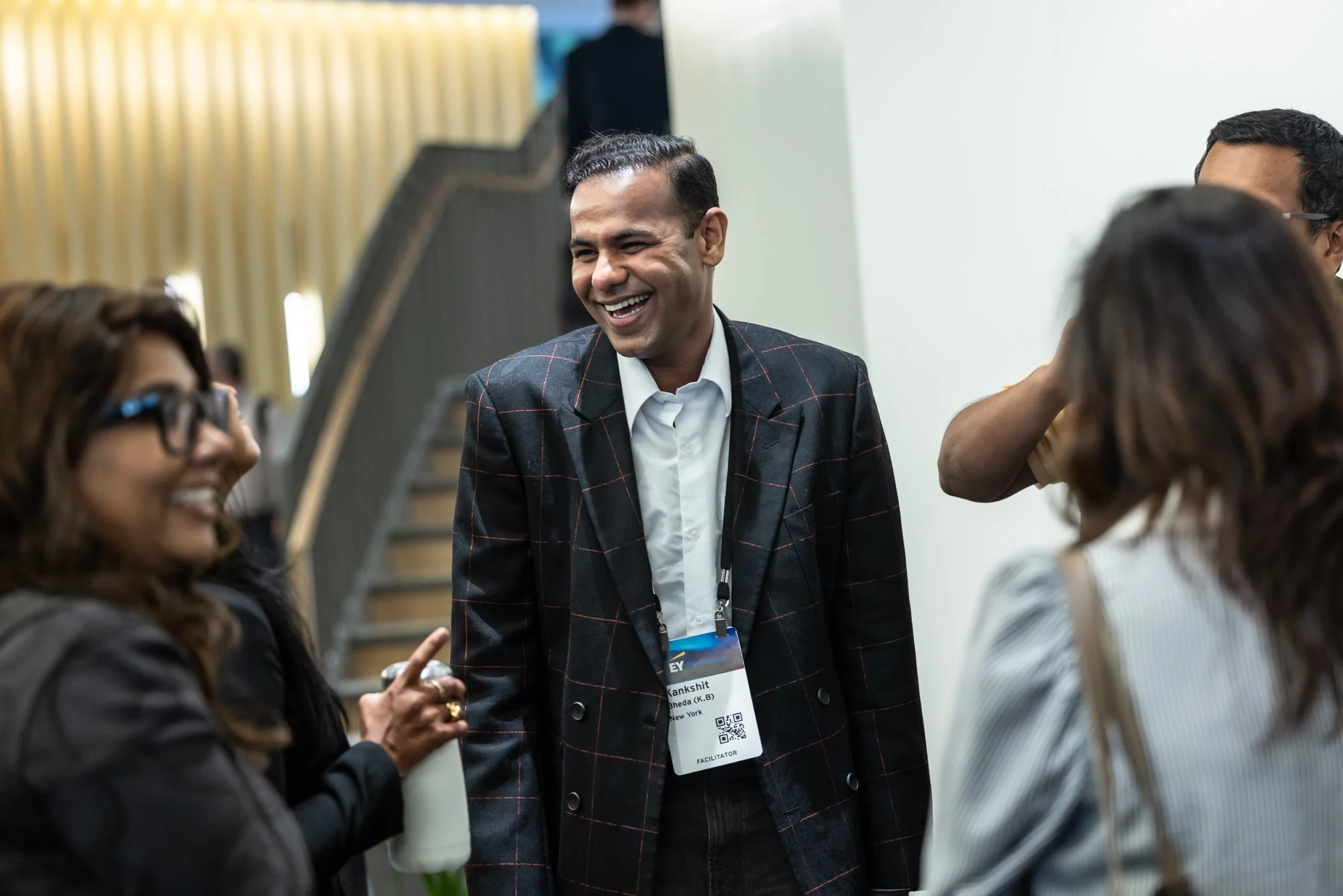 A group of diverse professionals in a business meeting, smiling and engaged in conversation, with a man in a suit and name badge at the center.