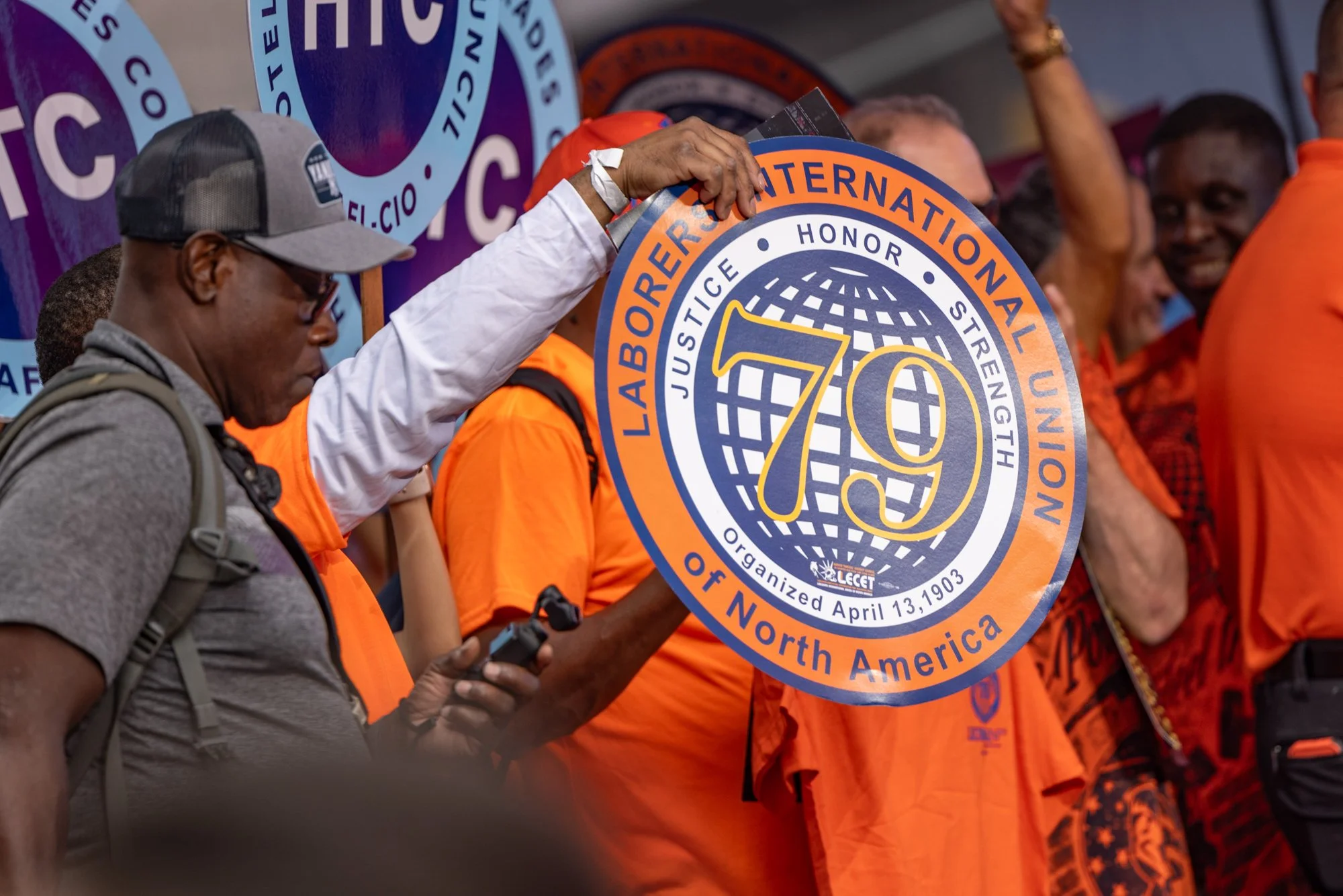People participating in a rally or march, holding a large circular sign with the text 'Judiciary International Labor Honor 70 North America' and a globe design. The participants are wearing orange shirts, and some are using their phones. One person i
