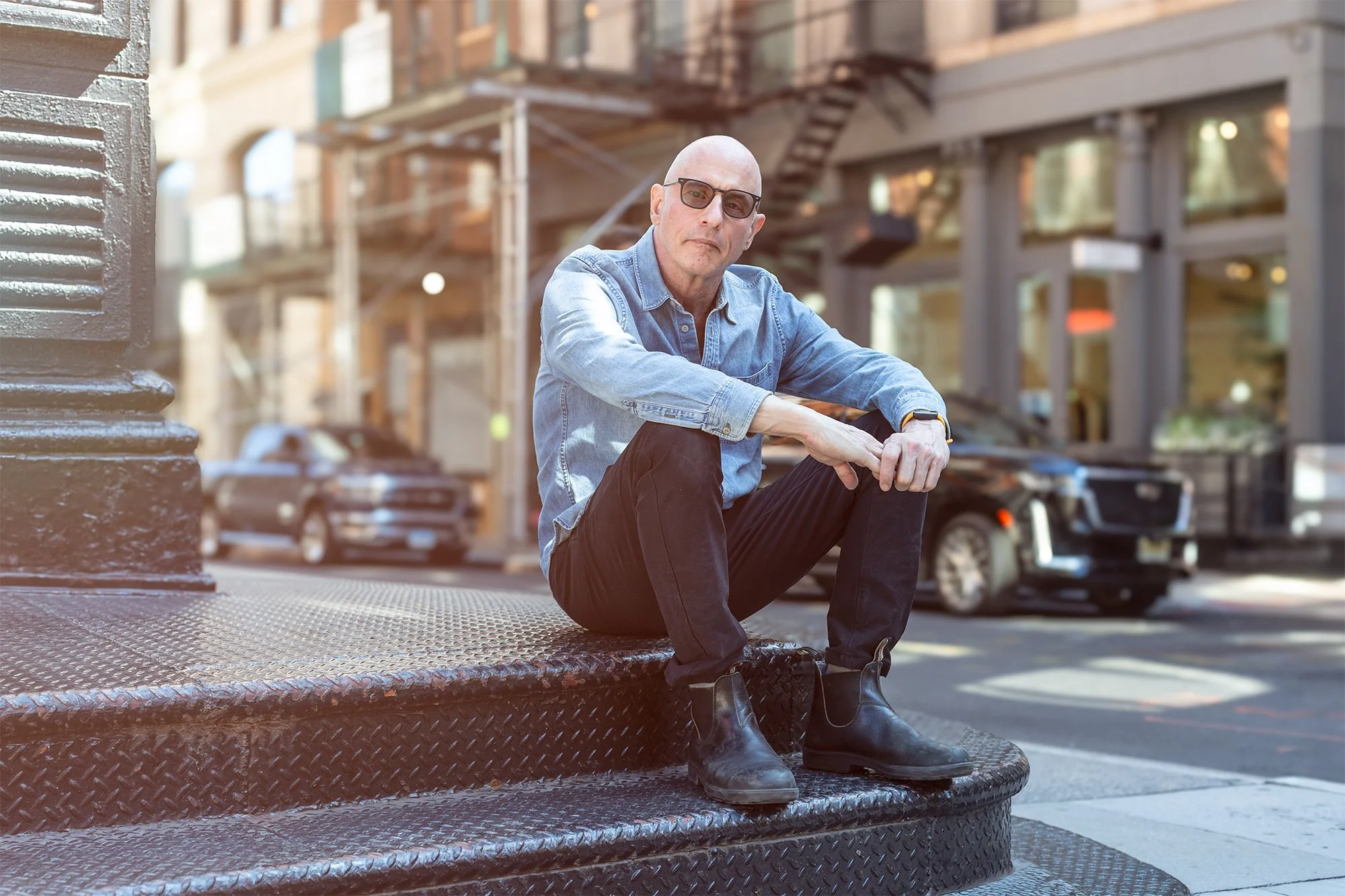 A bald man with glasses wearing a denim shirt and black pants, sitting on a black metal street corner, with buildings, cars, and a fire escape in the background.