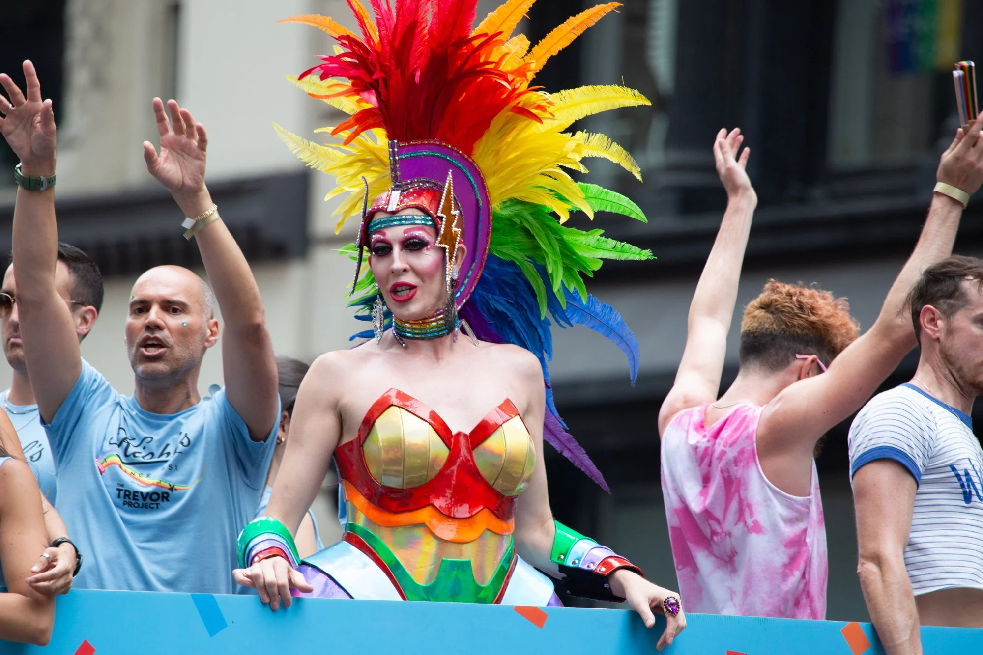 Drag performer in a colorful feathered headdress and jeweled costume celebrates with a crowd at a Pride parade.