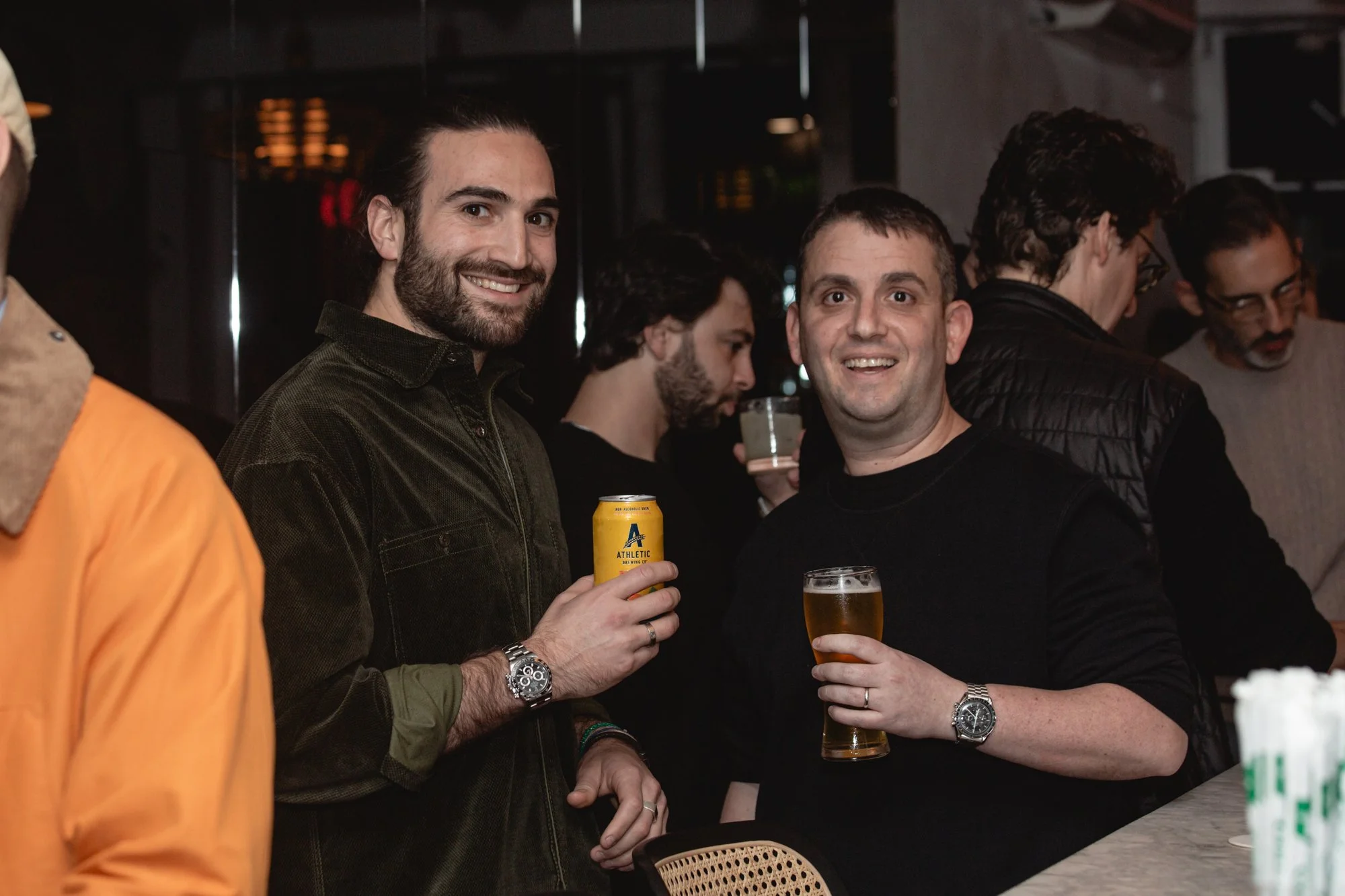 Two men smiling and holding glasses of beer at a bar or pub, with other people in the background.