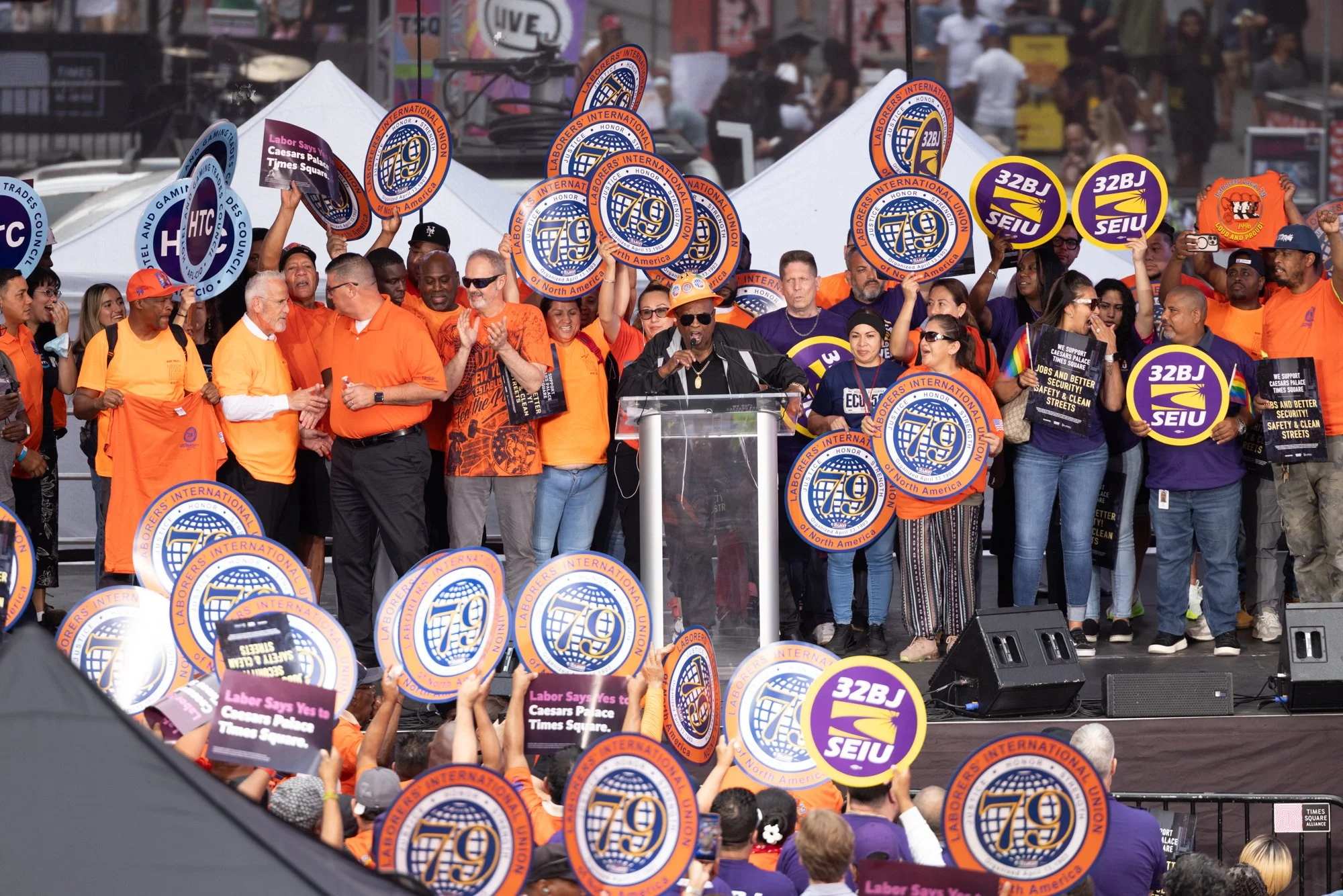A diverse group of protesters at a rally holding signs and banners with slogans. Some signs display logos of labor unions like SEIU. The crowd is gathered outdoors under tents, with many people wearing orange shirts and hats. A person at a podium app