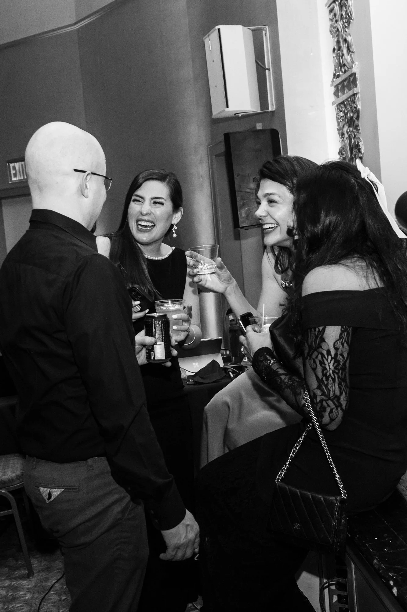 Four people gathered in a social setting, laughing and holding drinks, with two women in the center and two men on the sides, in an indoor environment.