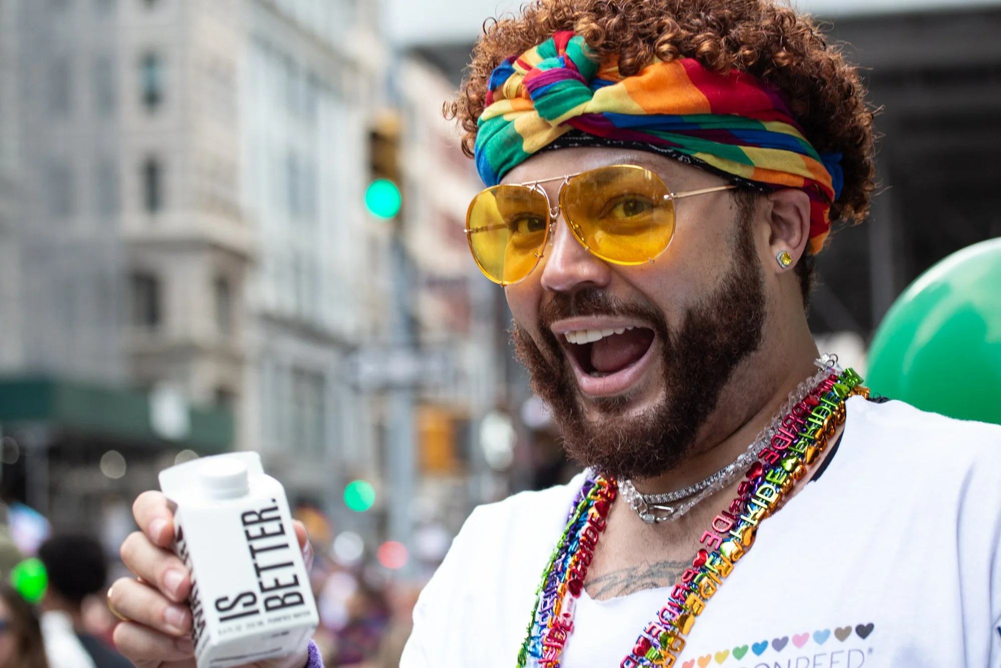 Smiling man wearing a rainbow headband, yellow sunglasses, colorful bead necklaces, and holding a white beverage can at a parade