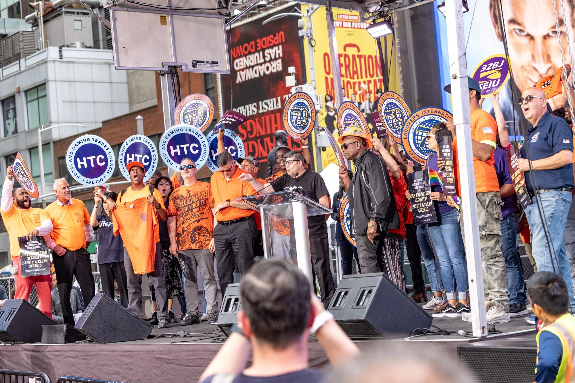 Crowd of people participating in a protest or rally on a city street, holding signs with union logos and slogans, with buildings and billboards in the background.
