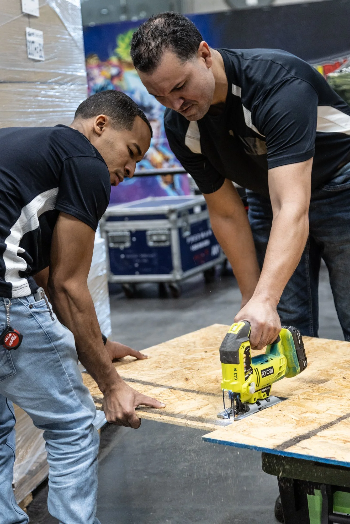 Two workers in black shirts measuring and cutting plywood with a Ryobi jigsaw at a workshop table.