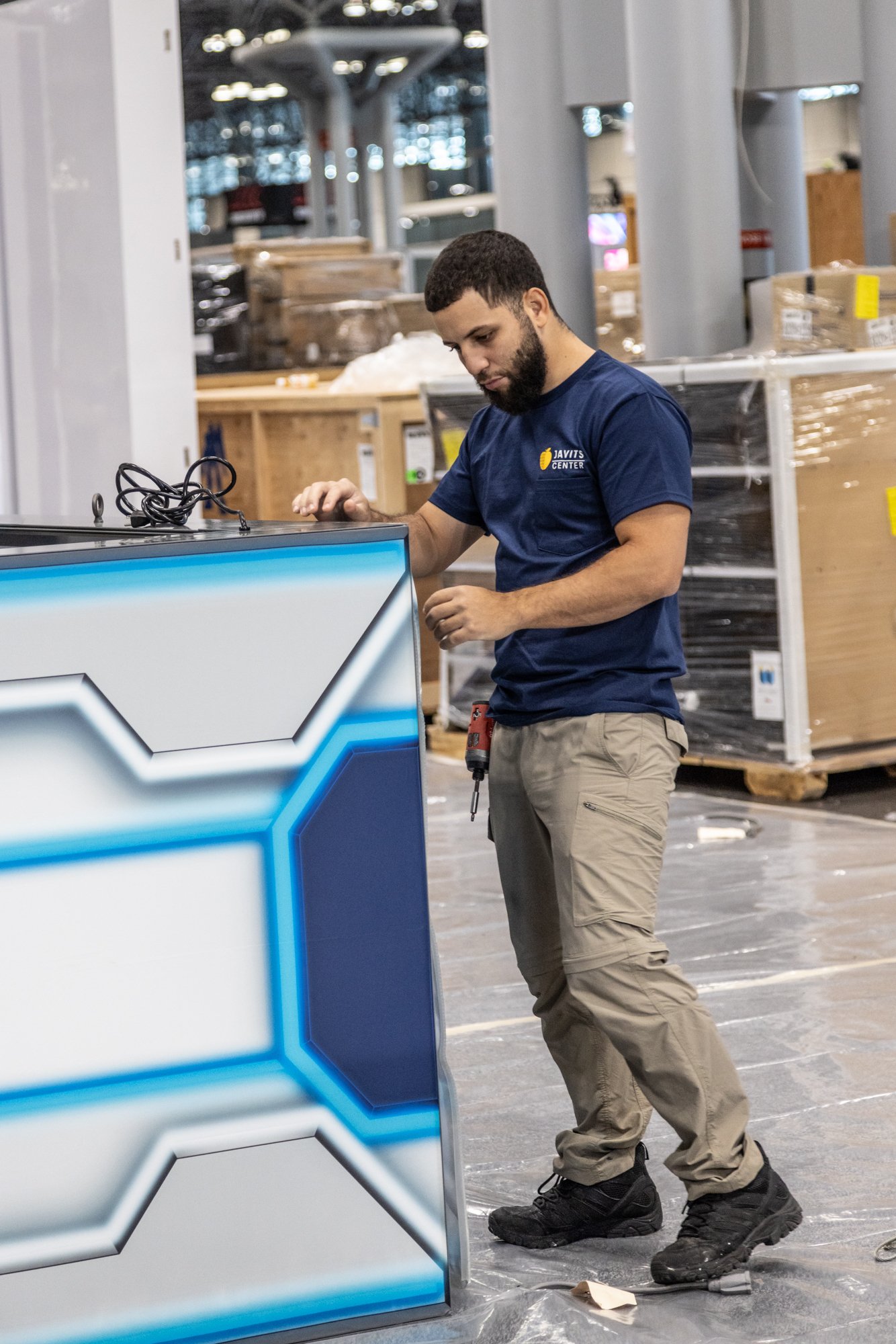 Technician in a Javit’s Center shirt assembling a blue-lit retail kiosk in a store.