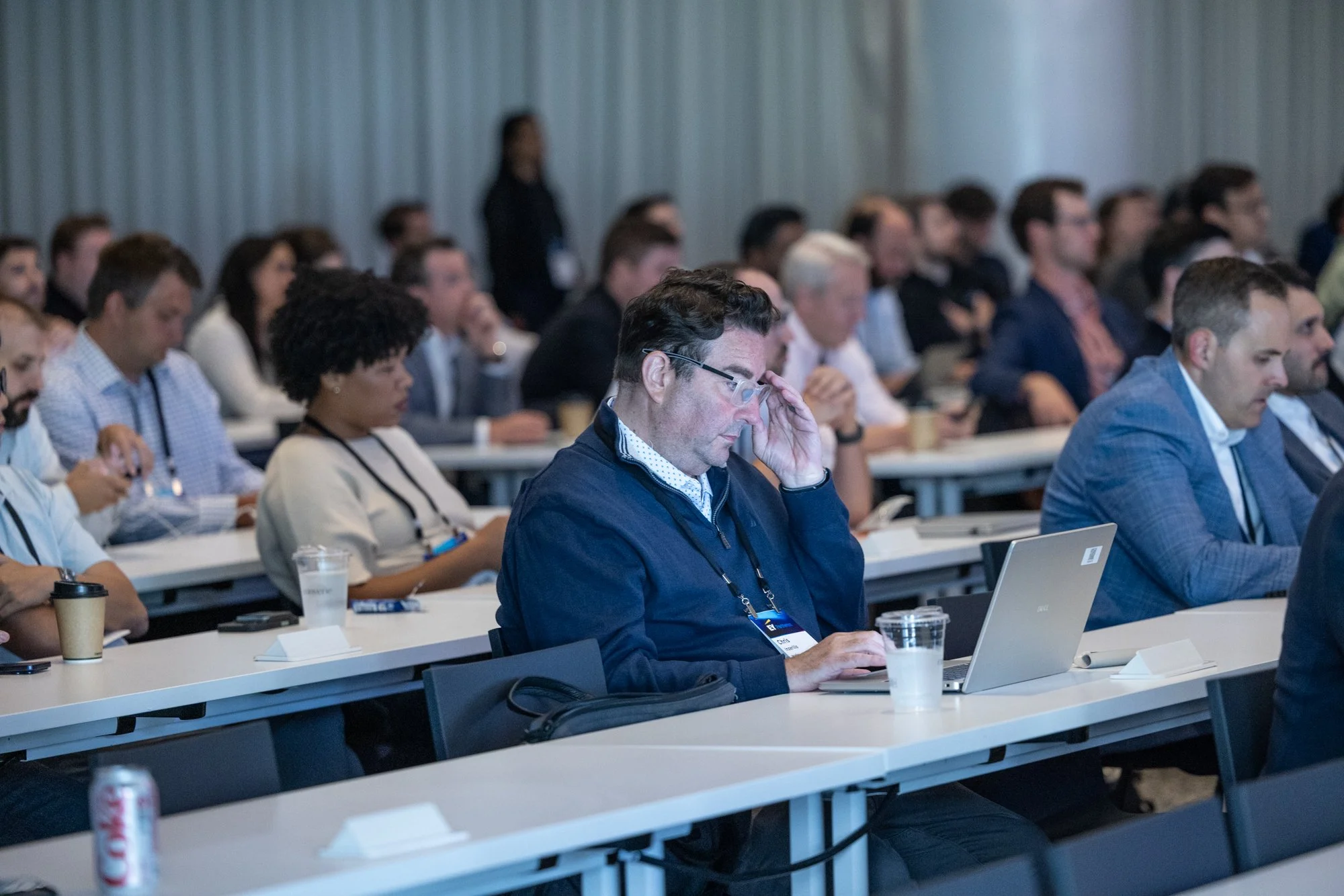 A diverse group of people attending a conference or seminar, sitting at long tables with laptops, notebooks, and drinks, listening to a speaker in a room with gray walls.