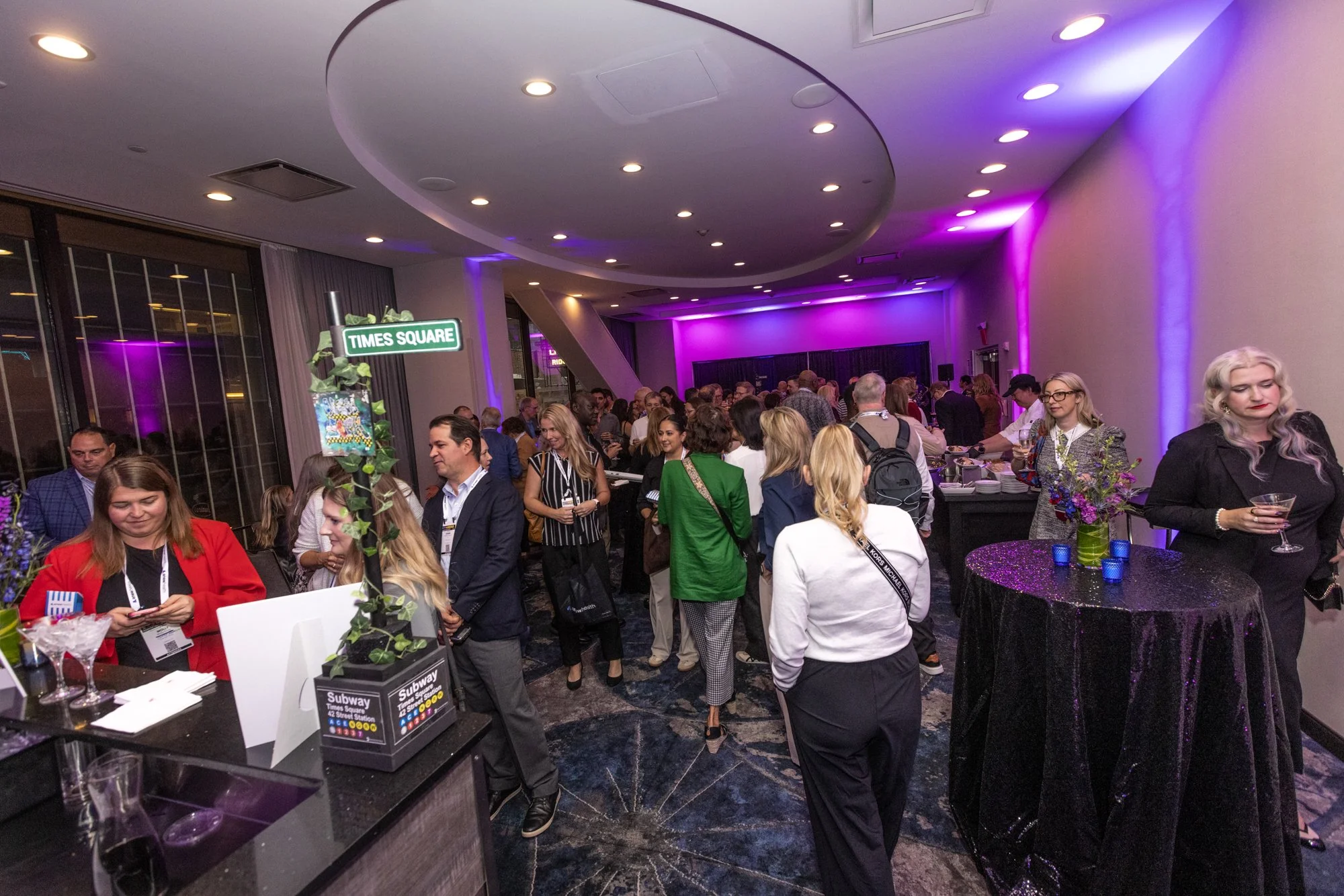 A crowded indoor event space with many people socializing and standing around tables decorated with purple flowers and candles. The room has a modern design with a curved ceiling and purple accent lighting. A green street sign reading 'Times Square' 